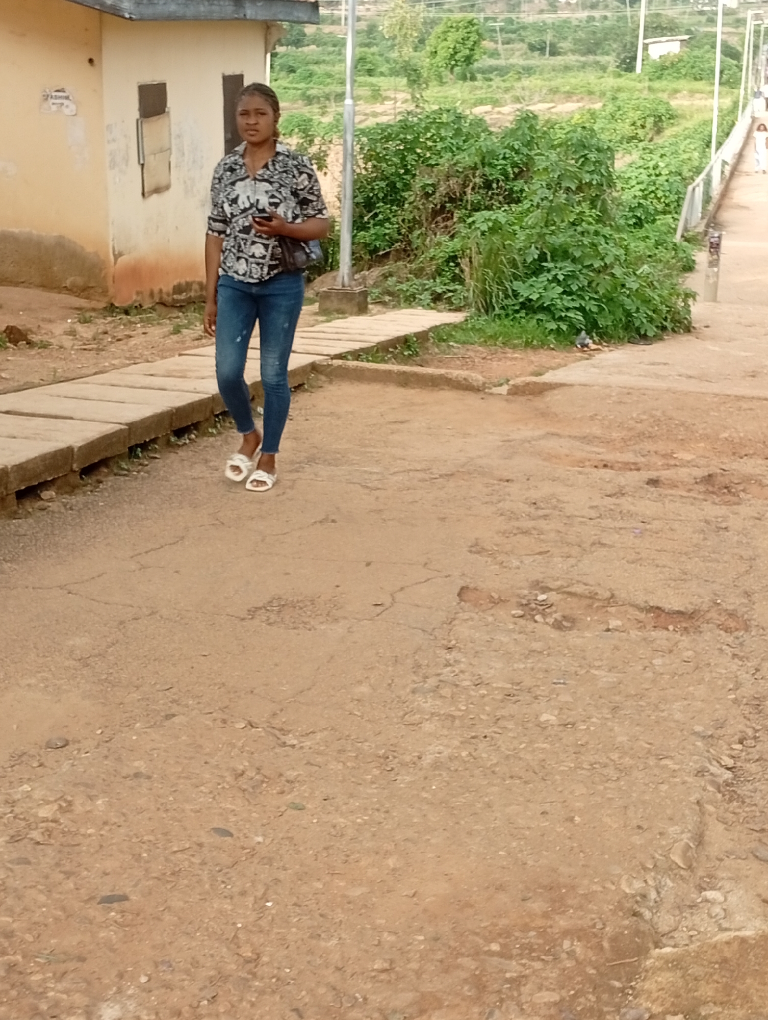 A young woman with dark hair pulled back is walking on a dirt path next to a concrete walkway. She is wearing a black and white patterned button-down shirt, blue jeans, and white slide sandals. She is carrying a dark purse over her left shoulder and holding a dark object, possibly a phone, in her right hand. She is looking forward as she walks. Behind her, to the left, is a light-colored building with two rectangular windows. To the right of the building and behind the woman is lush green vegetation and a sloped landscape. The ground is predominantly dirt with some cracked sections and scattered small stones. The scene appears to be an outdoor area in a moderately vegetated part of the city of Jos, Nigeria.
