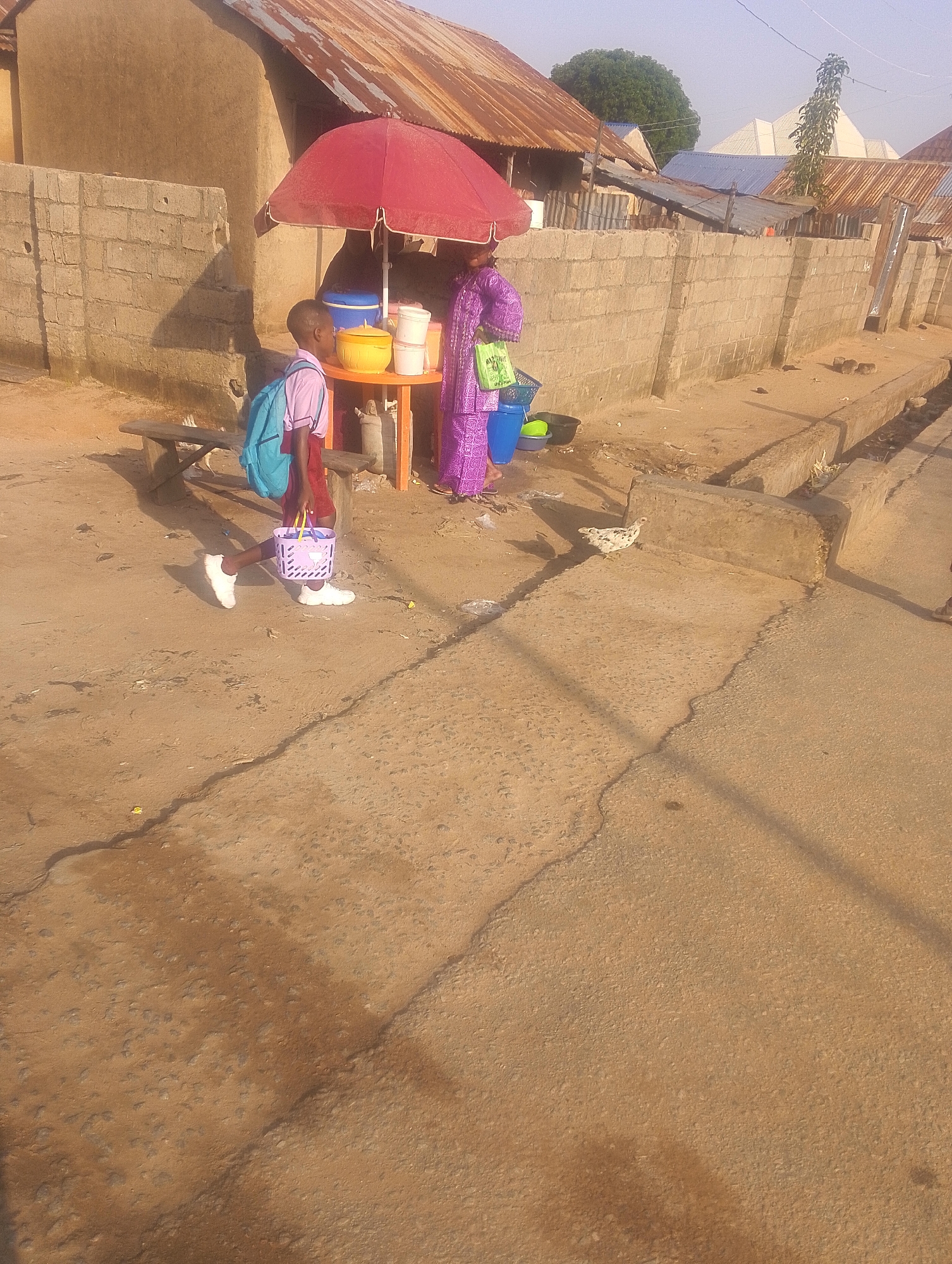 The image depicts an outdoor scene in Jalingo, Nigeria, likely on a street or market area. A young boy wearing a light blue backpack and a maroon skirt is walking, carrying a small pink basket. He is wearing white sneakers. Behind him, a woman in a purple patterned dress stands near a stall with a red umbrella. The stall displays various items including a yellow bowl, white containers, and blue buckets. A wooden bench is visible behind the boy, and a stone wall runs along the background. A chicken is present on the ground in the foreground. The environment appears to be dusty, with a concrete pathway and a dirt ground surface. The buildings in the background have corrugated metal roofs and mud-brick walls, typical of the region.