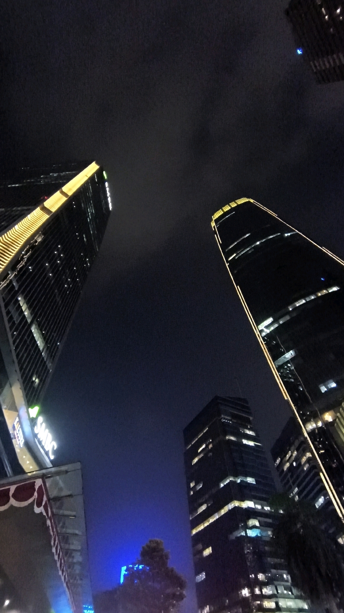 The image is a nighttime, low-angle shot of several modern skyscrapers in South Jakarta, Indonesia. The sky is dark and appears cloudy.
In the foreground and left of the image, a tall building with a facade illuminated by yellow lights runs vertically. Its lower section shows a portion of another building with a patterned awning. A sign with green lettering that appears to read "SMBC" is visible on this building.
To the right of the first building, a much taller, slender skyscraper dominates the central and right portions of the image. This building has a dark glass facade and is outlined by continuous yellow lights that accentuate its height and shape. Multiple windows on this skyscraper are lit from within, indicating occupancy.
In the middle ground, another, squatter skyscraper is visible. It also has a dark facade with numerous illuminated windows.
Further in the background and lower right, parts of other buildings with illuminated windows are visible.
At the bottom center of the image, a tree with dense foliage is silhouetted against a bright blue light emanating from a lower building or structure. The blue light appears to have the shape of a letter "F".
The overall scene depicts a vibrant urban nightscape with illuminated architectural structures, suggesting a bustling business or commercial district. There are no people visible in the image.