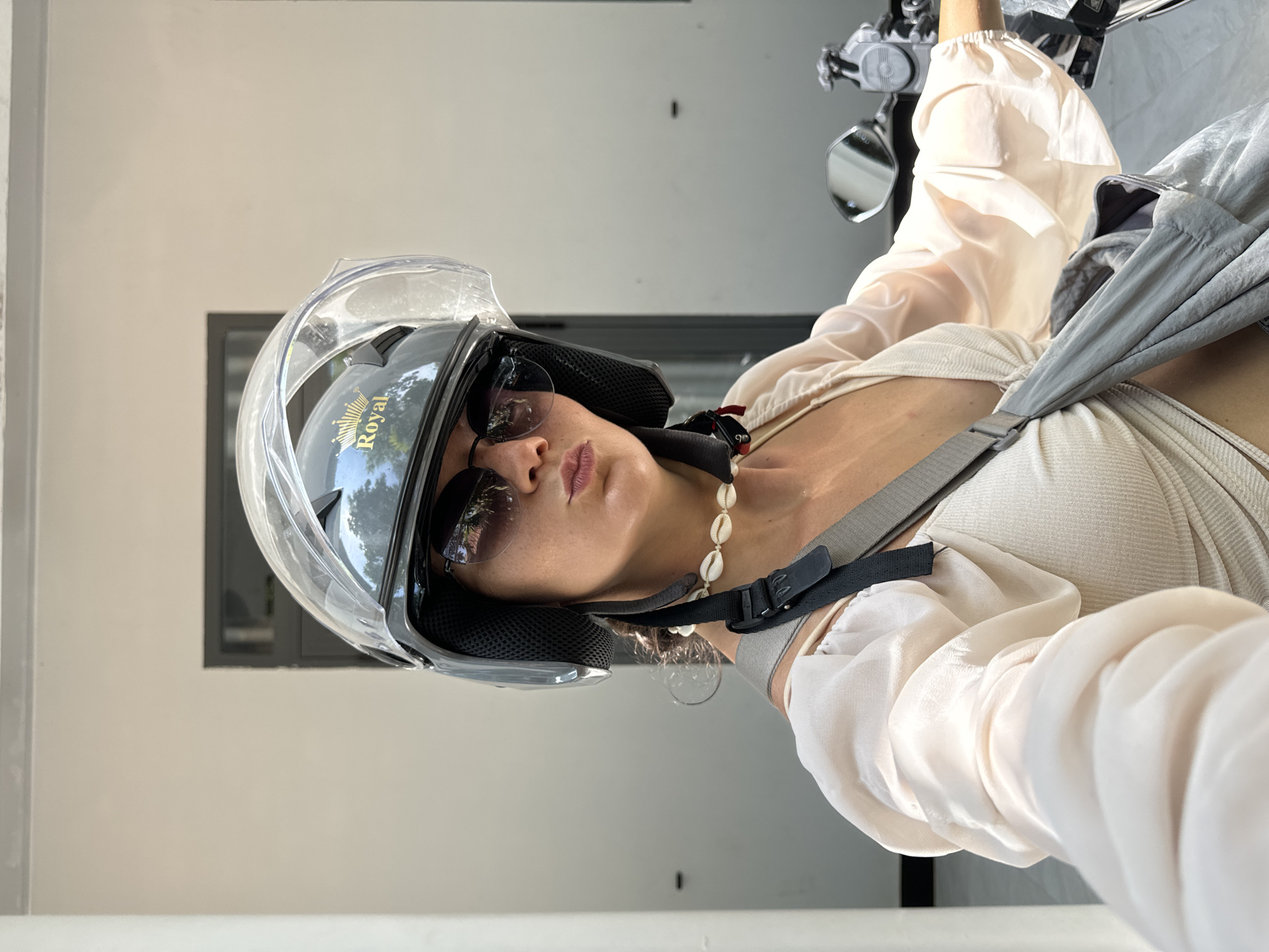 A young woman, wearing a helmet and sunglasses, is taking a selfie on a scooter in Mũi Né, Vietnam. The woman has her lips pursed for the photo, and she is wearing a light-colored top and a seashell necklace. A gray bag is slung over her shoulder. The background appears to be a white building with a dark-trimmed window. The sunlight suggests it is daytime.