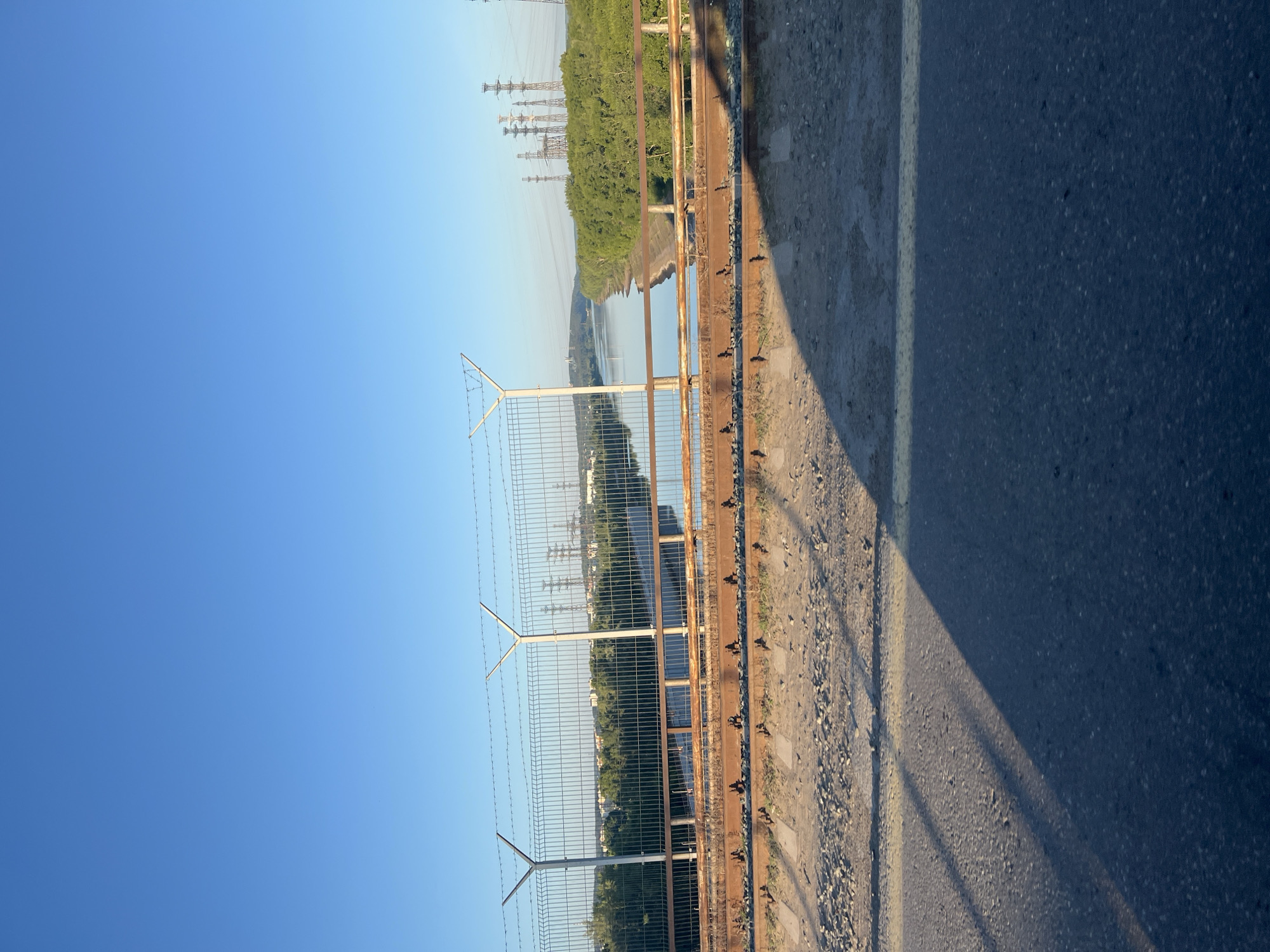 The image depicts a view from a bridge overlooking a wide river under a clear blue sky. The foreground features a segment of asphalt road with a yellow line, and beyond that, a dusty area with visible train tracks and rusty metal structures. A metal fence with barbed wire tops it, separating the bridge from the river. The river is calm and reflects the sky and the surrounding lush green trees that line its banks. In the distance, several power transmission towers are visible, along with a scattering of buildings, suggesting a populated area or industrial zone on the horizon. The overall scene is one of an industrial landscape bordering a natural waterway, likely taken during daylight hours. The presence of train tracks and industrial towers points towards the Chaikovsky, Russia area, known for its industrial development and proximity to the Kama River.