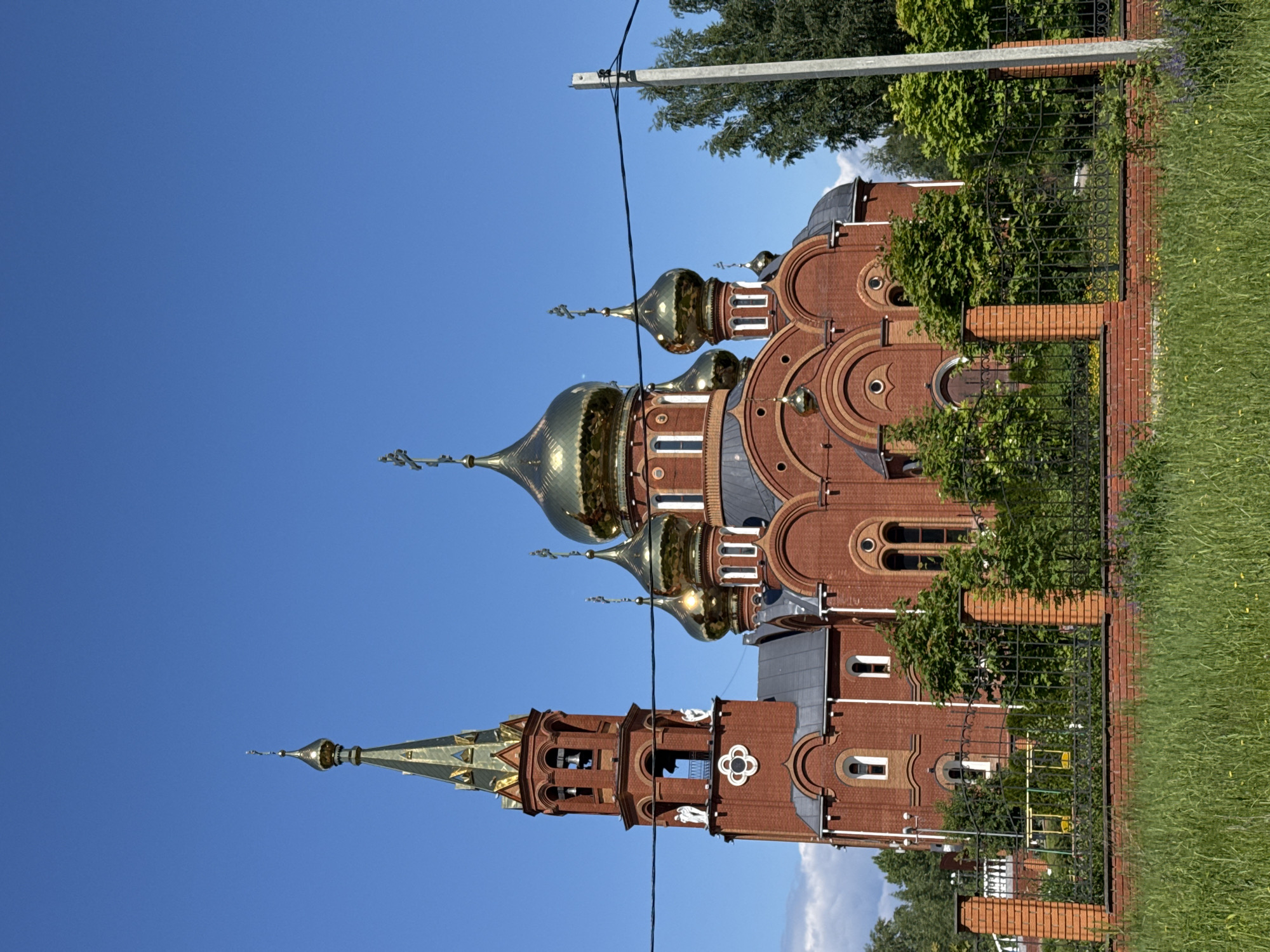 The image captures a large, ornate Eastern Orthodox church in Chaikovsky, Russia, bathed in bright daylight under a clear blue sky. The primary subject is the church, constructed from red brick, featuring multiple golden, onion-shaped domes, each surmounted by a cross. Architectural details include rounded arches above windows and decorative circular brickwork on the facade. To the left, a tall, slender bell tower rises, also made of red brick, crowned with a pointed golden spire and featuring arched openings.
The church grounds are bordered by a black metal fence with decorative scrollwork, supported by brick pillars. Lush green trees are visible around and behind the building, and green grass covers the foreground and extends to the right. A utility pole and a black power line cut horizontally across the upper part of the frame.
The scene is static, with no people or specific activities depicted. The clear blue sky and bright illumination indicate sunny daytime weather. There is no visible text in the image.