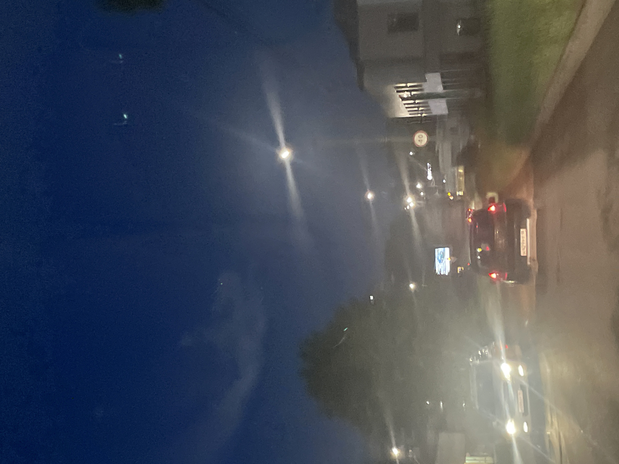The image depicts a nighttime scene on a street in Votkinsk, Russia. The sky is a deep blue, with faint clouds and what appear to be two distant lights or celestial bodies. In the foreground, a road is visible, with several vehicles. A white car is illuminated by its headlights, facing towards the viewer. To its right and further down the road, a darker colored car is visible, also with illuminated taillights. A speed limit sign indicating "40" is mounted on a pole. Buildings are present on the right side of the street, one appearing to be a light-colored building with multiple windows. Trees are also visible, with a large, shadowed tree prominently featured on the left side of the image. The lighting suggests it is dusk or nighttime, with artificial lights from streetlamps and vehicles contributing to the scene. The overall impression is of a quiet, possibly residential street at night.