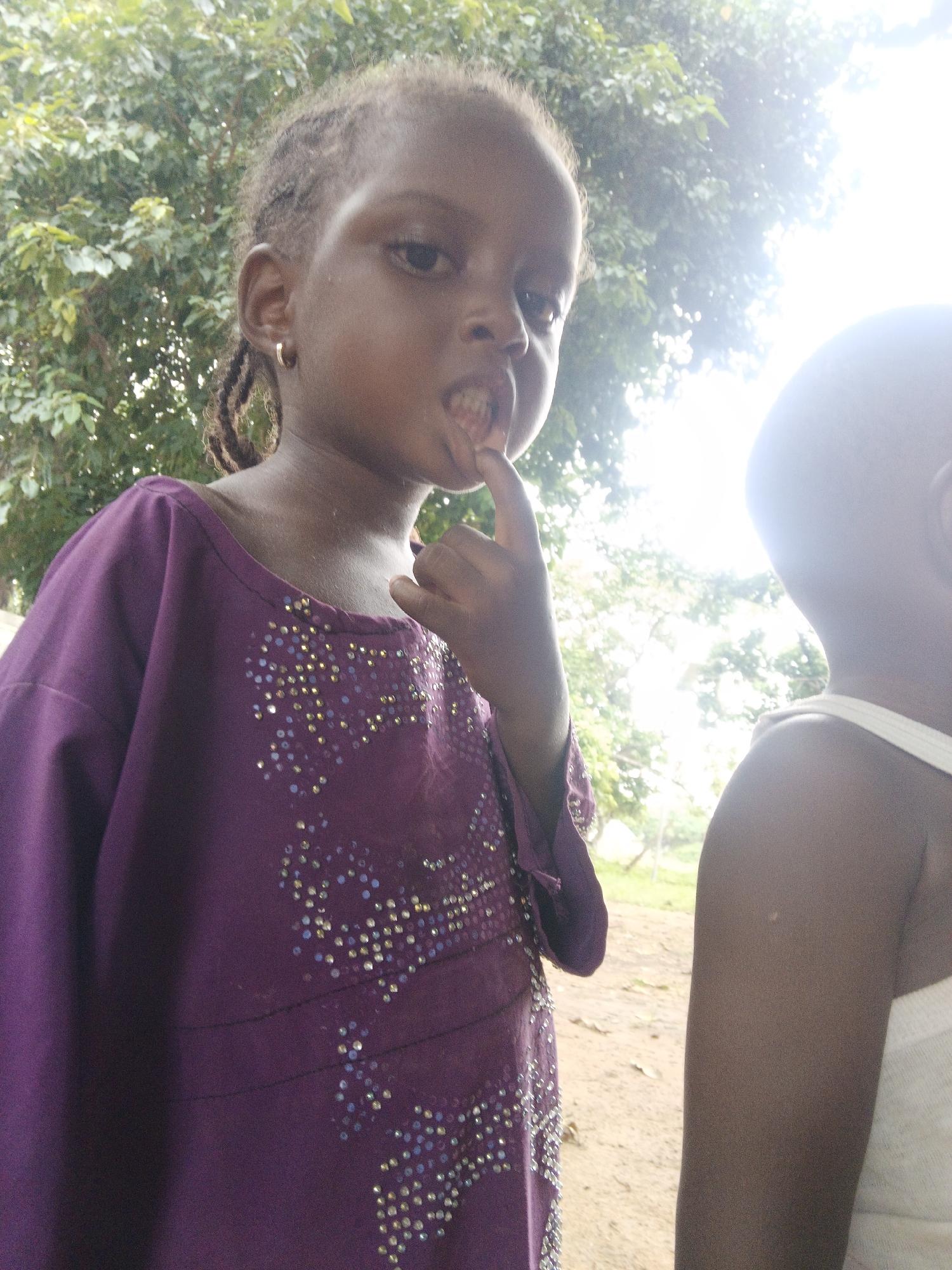 An outdoor, medium-shot, low-angle, natural light photograph shows two young Black children standing side-by-side, with a blurry background of green trees and bright sky. The child on the left, a girl, is in the foreground, facing the camera at a three-quarter angle. She has dark skin, dark hair in braids, and is wearing a purple dress with beaded embellishments. Her right index finger is raised to her mouth, and she appears to be biting her nail. She is wearing a gold hoop earring in her left ear. To the right of the girl, the back of the head of another child, likely a boy, is visible. This child is wearing a white tank top. The ground is visible in the lower part of the image and appears to be dirt and leaves.