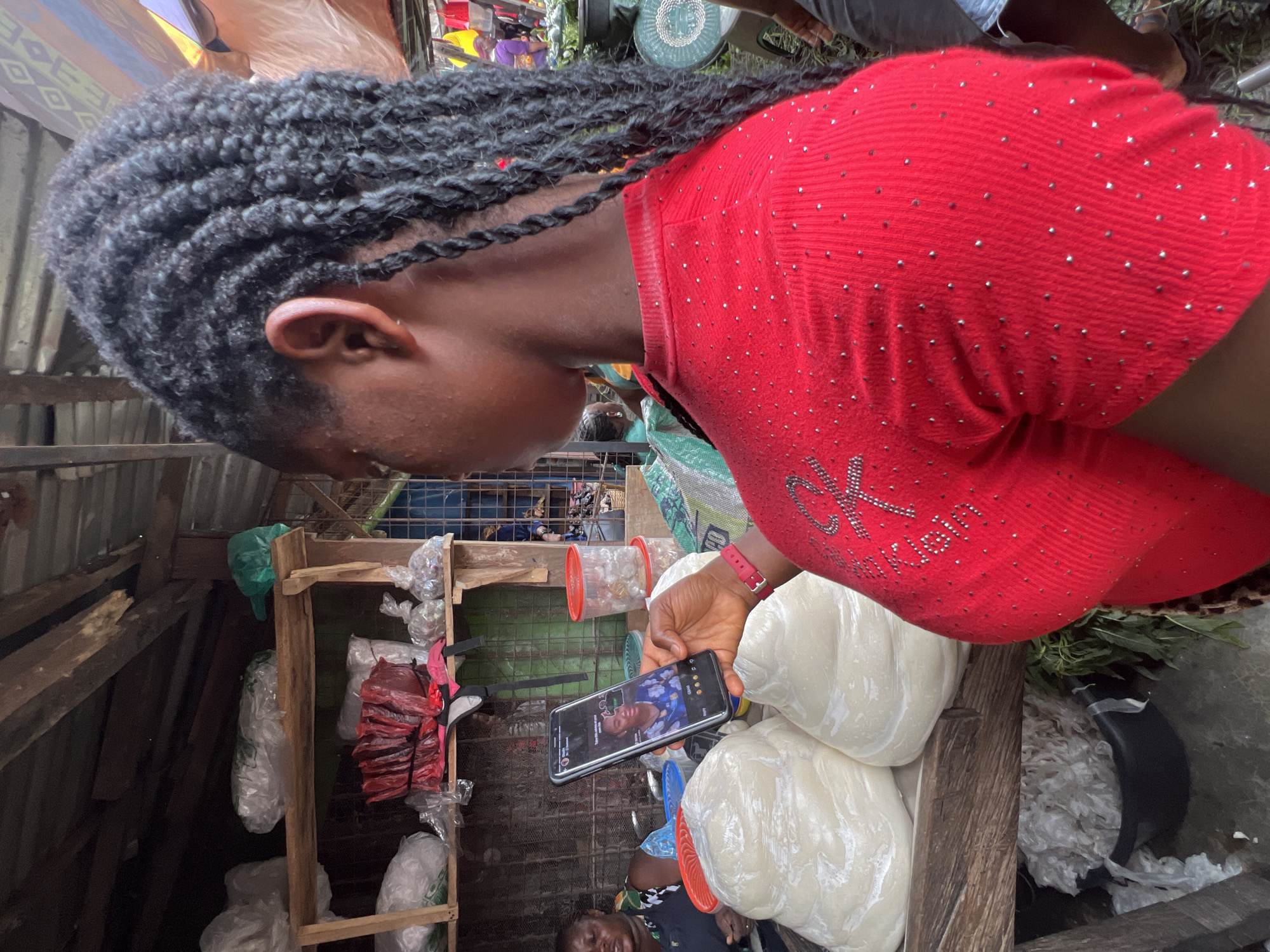 In Nkpor, Nigeria, a young woman with braided hair is holding a smartphone, displaying a video call with a woman in a blue dress. She is standing in an open-air market, surrounded by produce and goods. Large, white, plastic-wrapped food items are stacked on a wooden table in front of her. Shelves behind her are stocked with more items, some in plastic bags. Another woman, wearing a black and white patterned top, is visible in the background, seated behind the table. The scene is typical of a bustling marketplace, with a variety of goods and people engaged in commerce.