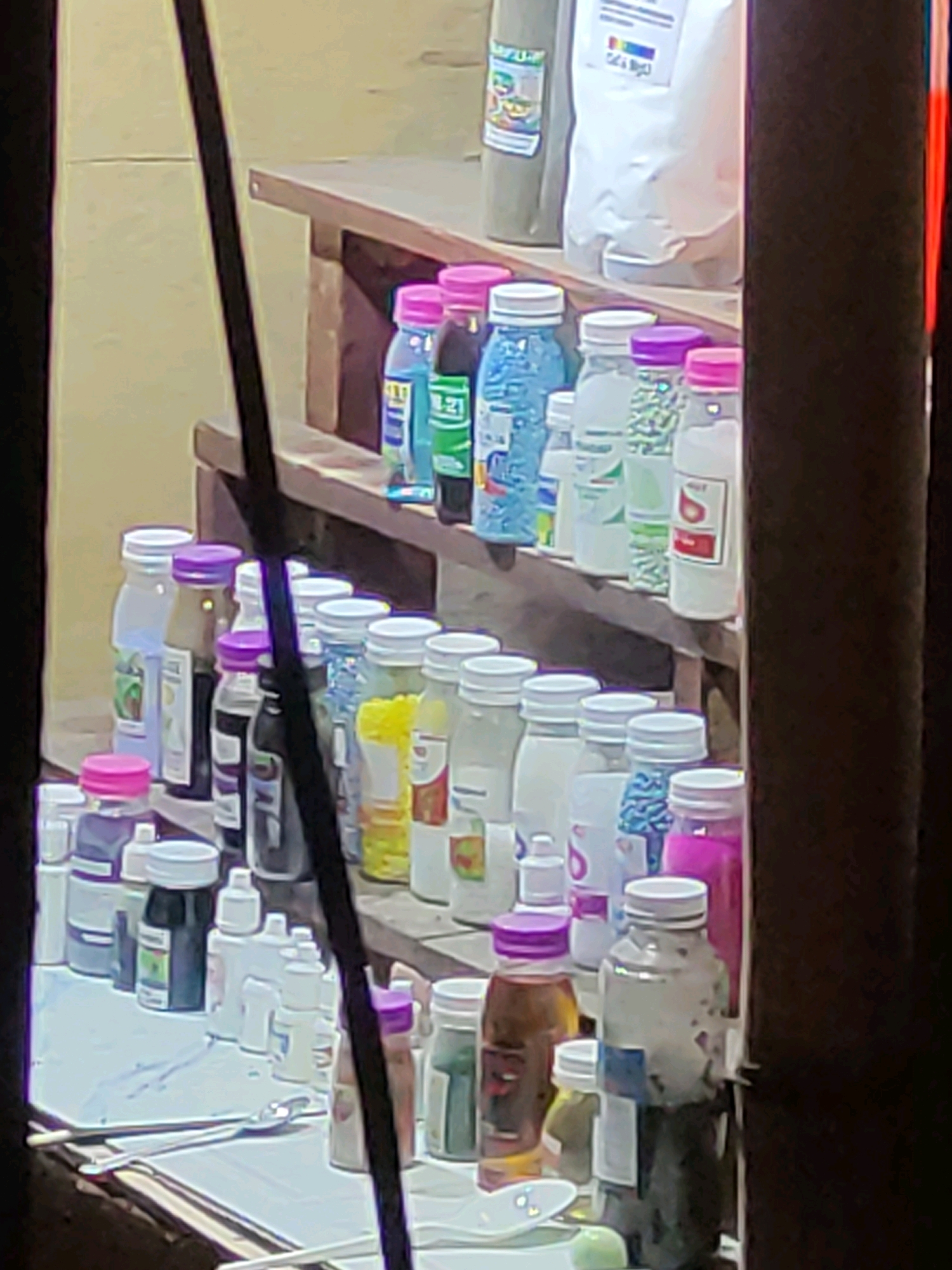 The image depicts a shelf filled with various bottles of liquids and powders. The items are arranged in rows on wooden shelves. Some bottles are clear, while others are colored. Many have white caps, but there are also some with pink, purple, and blue caps. Labels are visible on some of the bottles, but the text is largely illegible due to the image quality and distance. In the foreground, a white surface with some utensils, possibly spoons, is visible. The background appears to be a wall or structure. The overall impression is that of a retail display, likely in a small shop or market stall. The scene is located in Tiudan, Indonesia, as indicated by the prompt.