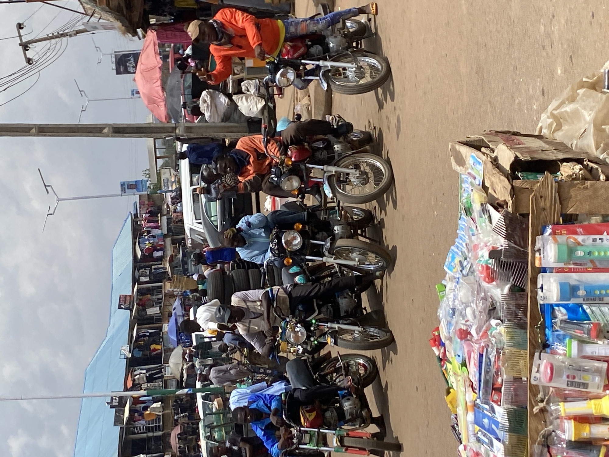 This image depicts a bustling street scene in Bukuru, Nigeria. A line of motorcycles is parked along the right side of the road, each with a rider on it. The riders appear to be waiting or resting. A vendor's stall laden with items like combs and flashlights sits beside the motorcycles. Across the street, numerous stalls with vendors and shoppers are visible, indicating a busy marketplace. The sky is overcast, and power lines crisscross above the street. Umbrellas provide shade for some vendors and shoppers. The overall impression is of a vibrant, crowded commercial area.
