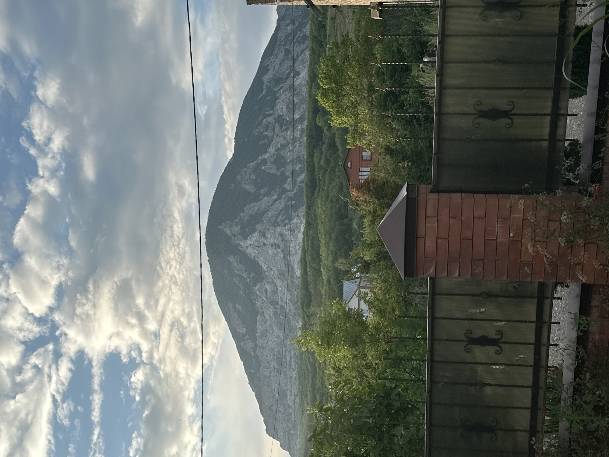 The image shows a natural landscape with a large mountain in the background. The mountain has a rugged, rocky surface. The foreground features a green area with trees, and a building, possibly a house, is visible amidst the foliage. A fence, made of metal and translucent material, is prominent in the right side of the image. The sky is partly cloudy, with some bright areas suggesting it might be daytime. A thin wire stretches across the frame from the top left. The overall scene appears to be a rural or semi-rural setting in a mountainous region. There is no visible text in the image. The orientation of the image suggests it may have been taken on its side.