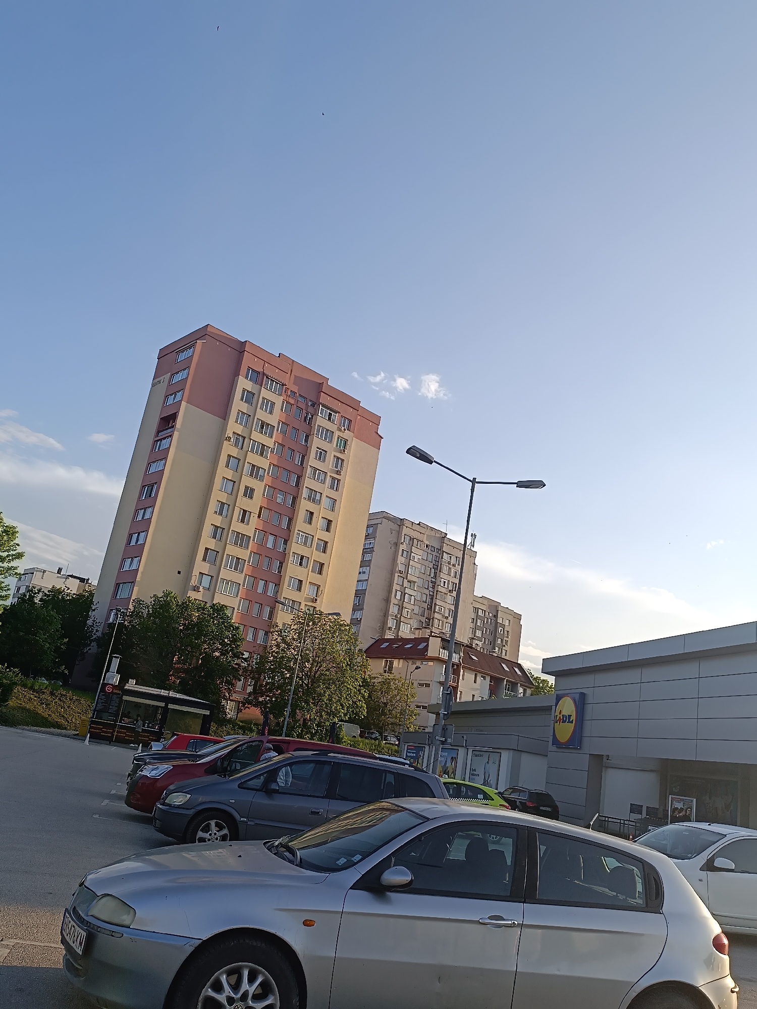 The image shows a view in Pleven, Bulgaria, featuring a Lidl supermarket with its yellow and blue logo visible on the building's facade. A parking lot in front of the store contains several parked cars, including a silver hatchback in the foreground. In the background are tall residential buildings with beige and pink exteriors. A street lamp stands near the Lidl building. The sky is blue with scattered clouds.