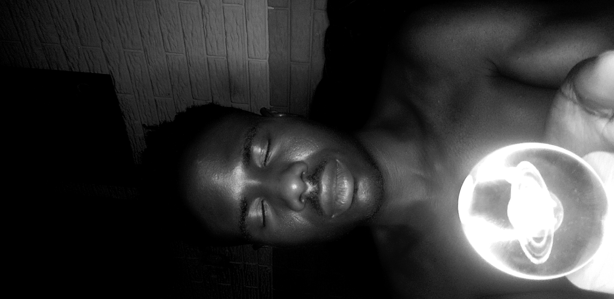 The image is a close-up, high-contrast black and white photograph of a Black man lying on his side. The man has his eyes closed, short hair, and a mustache. The lighting is dramatic, with a bright light source emanating from a circular object held near his face. The background consists of a textured wall resembling brick or tile. The location is Narayi, Nigeria. There are no clear interactions or actions visible.