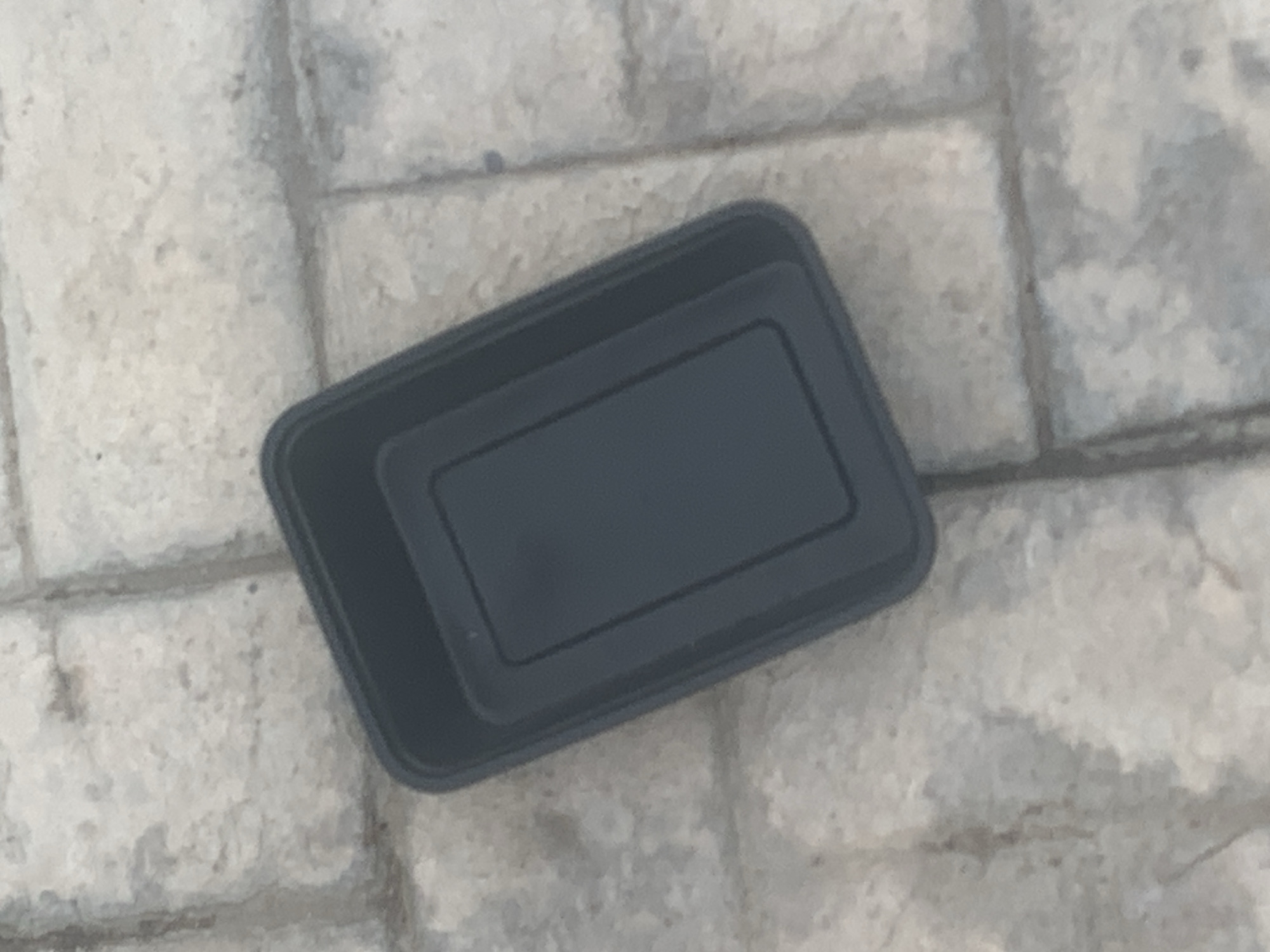 A dark gray, rectangular plastic food container is resting on a light gray, rectangular paved surface. The container is empty and appears to be on its side with its opening facing towards the top left of the frame. The paved surface is composed of individual stone-like blocks, with visible grout lines. The lighting suggests an outdoor environment. The image does not contain any people or specific landmarks that would allow for precise location identification in Jalingo, Nigeria.