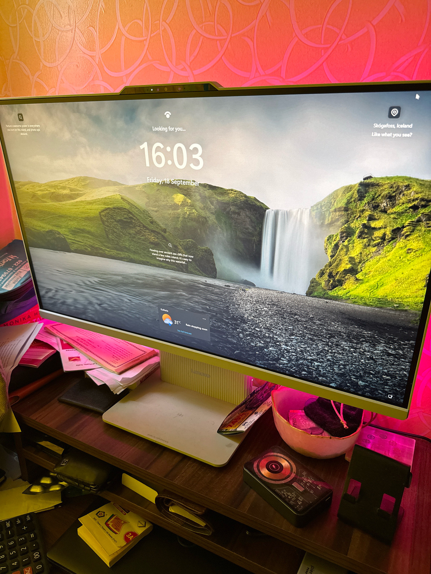 The image displays a computer monitor with a lock screen that shows a waterfall in Iceland. The time is 16:03 on Friday, September 18th. The location "Skógafoss, Iceland" is visible. Below the waterfall image, there's a weather forecast for Kolkata, India, indicating a temperature of 31°C with rain. The monitor is positioned on a wooden desk cluttered with various items, including papers, a calculator, a wallet, and a phone stand. The desk area is illuminated by pink LED lights.