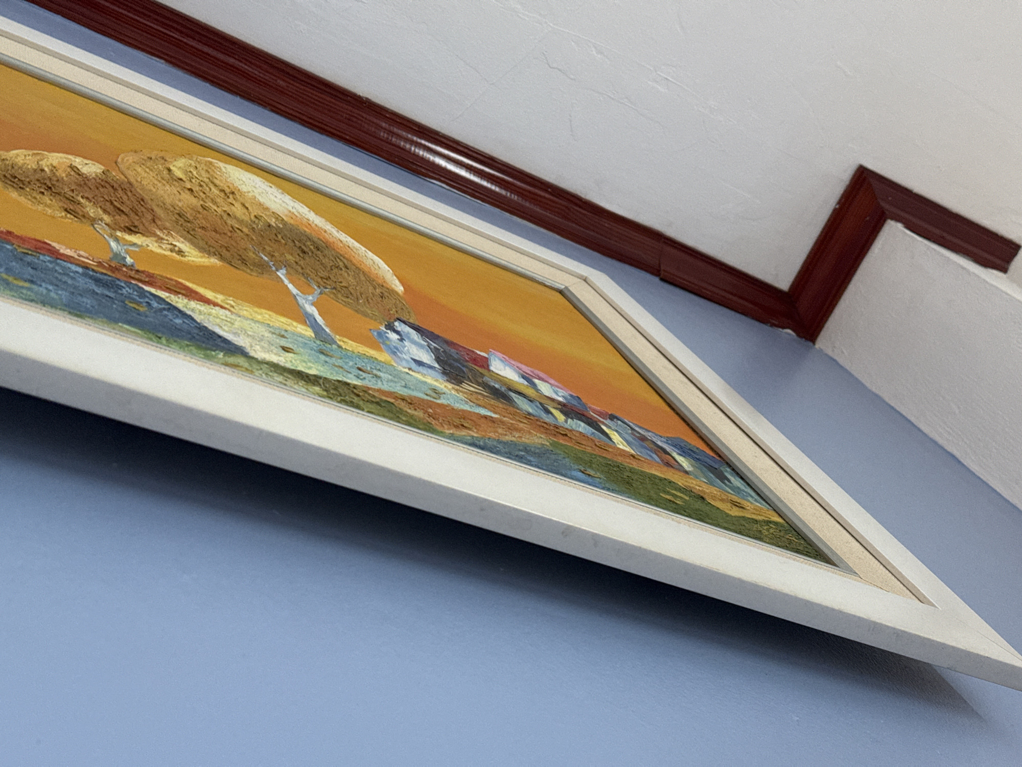 The image shows a close-up, oblique view of a framed painting on a wall. The painting features abstract, textured brushstrokes in vibrant colors, primarily orange, yellow, blue, and green, suggesting a possible depiction of natural elements. The painting is housed in a white frame, with an additional dark red trim framing the edge of the wall. The wall itself is painted a light blue, and the area to the right of the red trim is a textured white surface, possibly a ceiling or another wall. The lighting in the scene casts a soft shadow from the painting onto the blue wall. The scene appears to be indoors, with no exterior elements visible. The specific location in Hà Nội, Vietnam cannot be determined from the visual information present in the image.