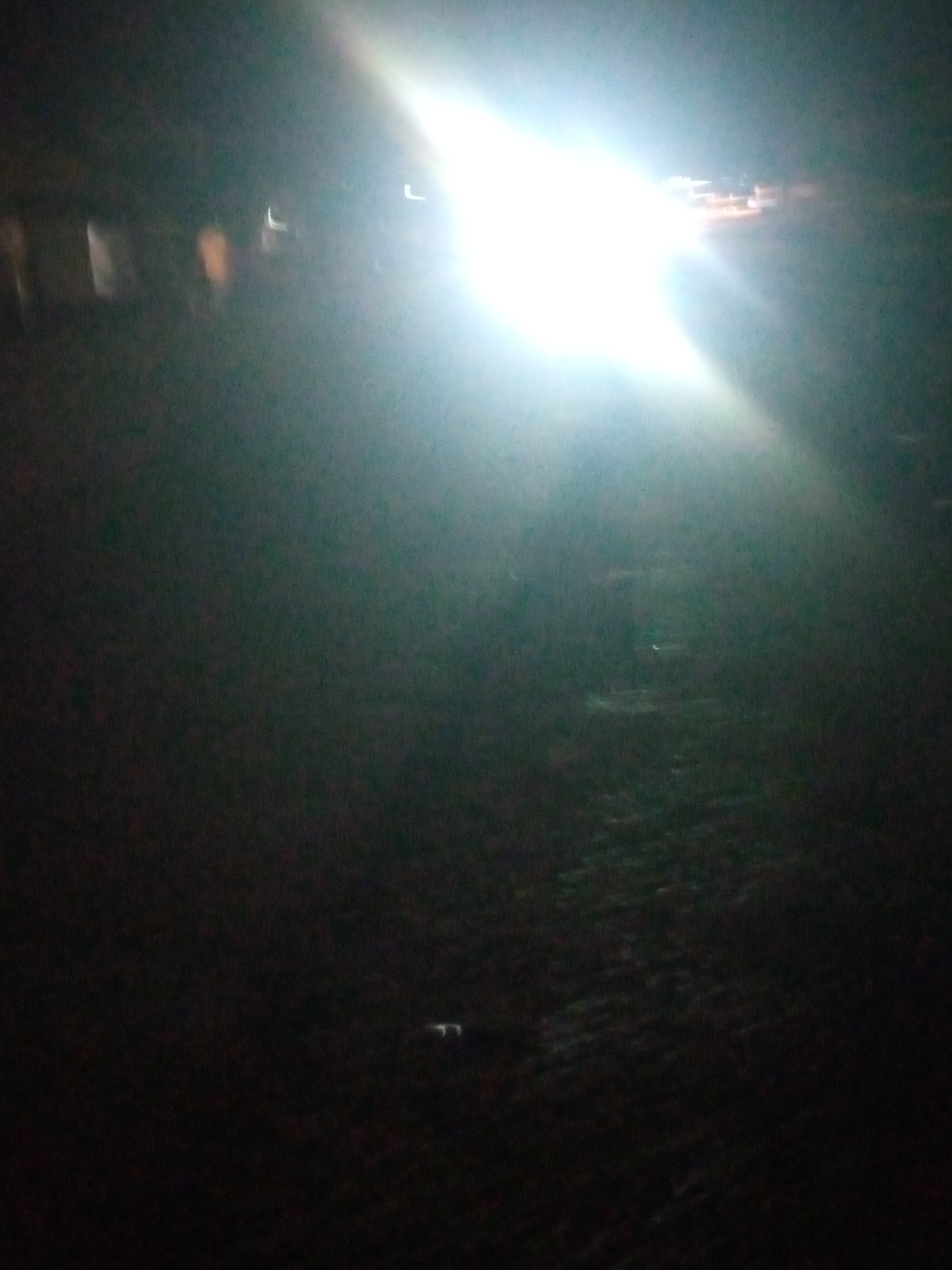 The image depicts a dimly lit outdoor scene, likely at night, with a bright light source dominating the upper right portion of the frame. The light appears to be from vehicle headlights or a strong artificial light, casting a glare and lens flare across the image. The ground surface is visible in the lower portion of the image and appears to be dark and possibly paved or covered in gravel, with some lighter patches suggesting reflections or small objects. In the upper left quadrant, there are faint, blurred shapes that could be buildings or structures, illuminated by ambient light or reflections from the primary light source. The overall impression is one of low visibility and a potentially urban or semi-urban environment at night. The image does not provide specific details that would allow for definitive identification of the location within Jalingo, Nigeria, beyond it being an outdoor setting with artificial lighting.