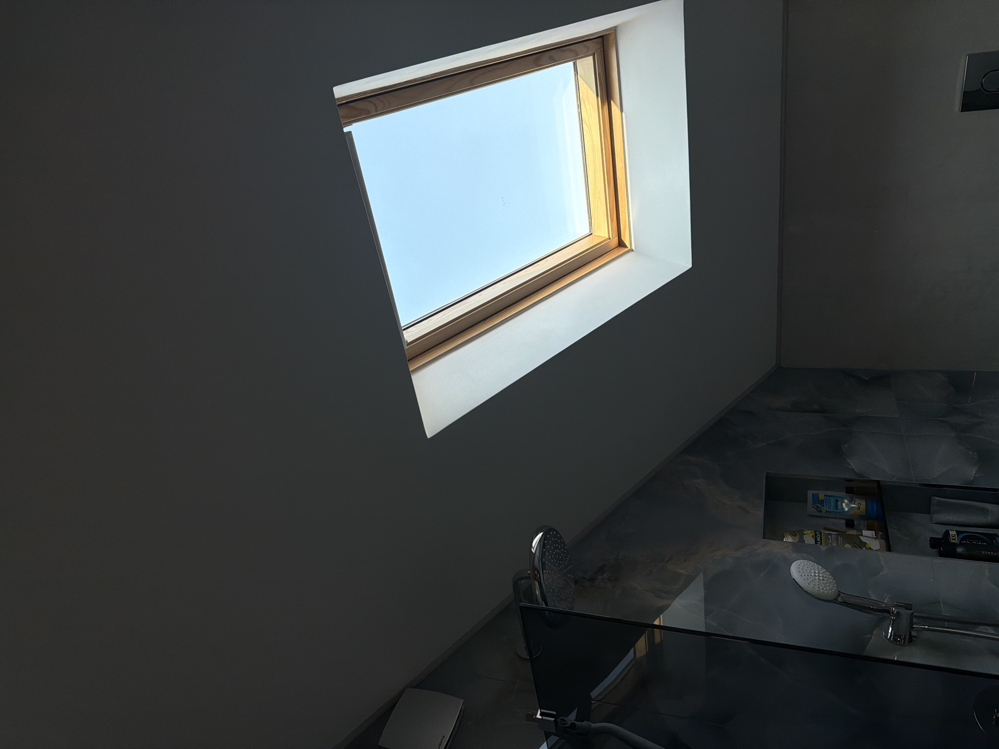 The image shows the interior of a bathroom in Buhovo, Bulgaria. A skylight is visible in the ceiling, framing a view of the blue sky. Below the skylight, the scene transitions to a shower area with dark marble-like tiles, a recessed shelf holding toiletries, and a shower head. A glass panel partially encloses the shower area. A toilet is visible in the corner of the room.