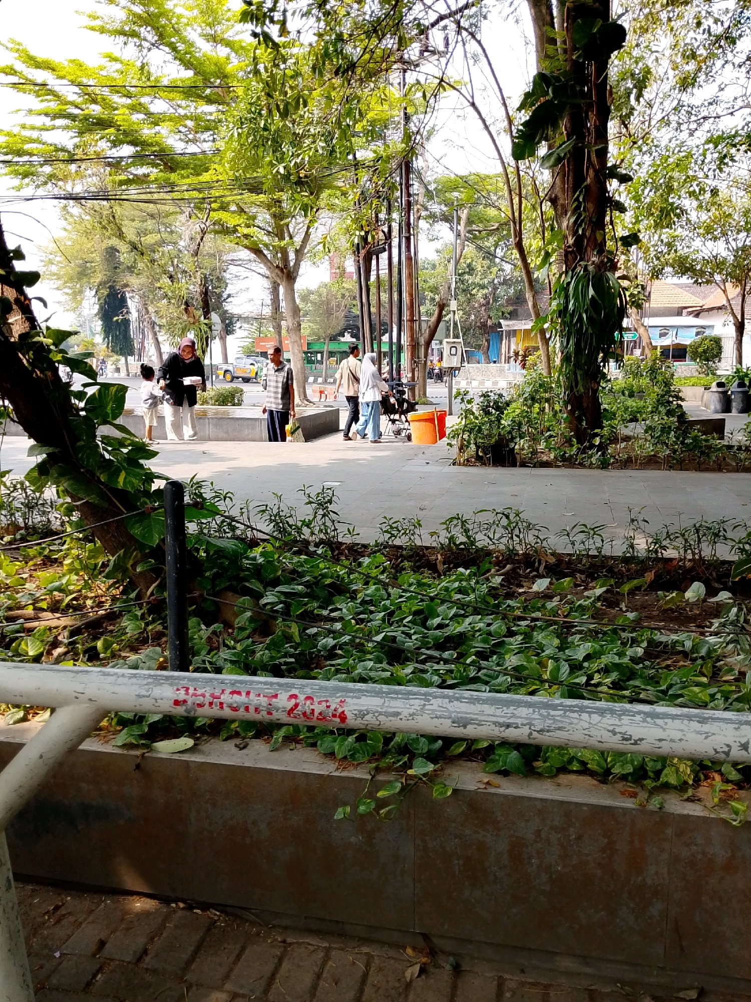 The image depicts an outdoor scene with people walking along a paved walkway lined with trees and greenery. In the foreground, a metal railing with red graffiti that reads "2020" is visible. Behind the railing, a concrete planter box holds numerous small plants. The walkway is bordered by a sidewalk. Several individuals are present, including a woman in a headscarf holding a white box and a child in a white shirt and shorts. Further down the walkway, a man in a striped shirt and another person in a white headscarf and blue pants are walking. In the background, there are more trees, buildings, and street infrastructure, suggesting an urban environment. The overall setting appears to be a public park or a pedestrian area within Kediri City, Indonesia, characterized by a sunny day with abundant foliage.