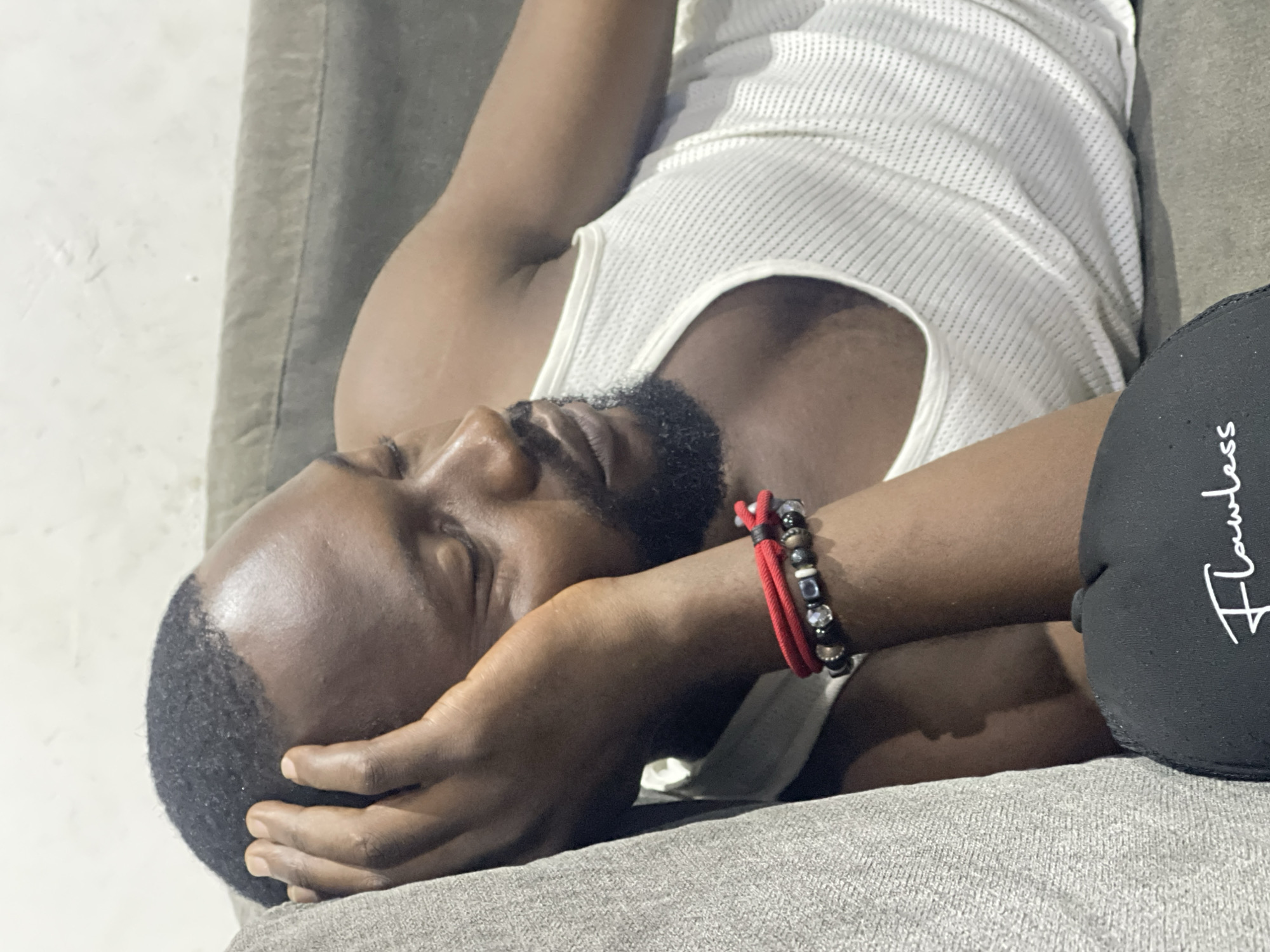 A man is lying on his side on a couch, his eyes closed and his head resting on his arm, suggesting he is sleeping or resting. He is wearing a white ribbed tank top and has a beard. A black fabric item with white text is visible on his right side. On his left wrist, he wears a bracelet made of red cord and various beads. The setting appears to be indoors, with a couch and a textured wall visible in the background. The lighting suggests it might be evening or night, or that the room is dimly lit. The overall mood is one of relaxation and repose. The visible text on the black fabric reads "Flawless". The location context provided is Abuja, Nigeria.