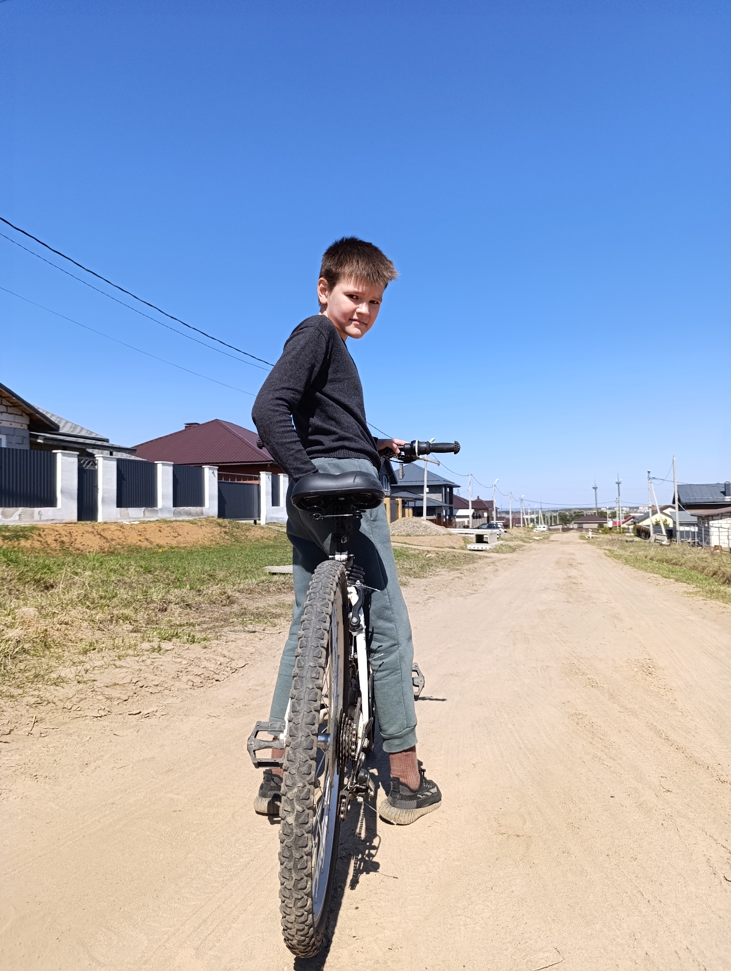 A young boy stands on a bicycle on a dirt road in Chaikovsky, Russia. The boy, dressed in dark clothing, is facing away from the camera but turning his head to look back. He is positioned on the bicycle with his feet on the pedals. The background shows a neighborhood with houses, a dirt road, and a clear blue sky. Power lines are visible overhead.