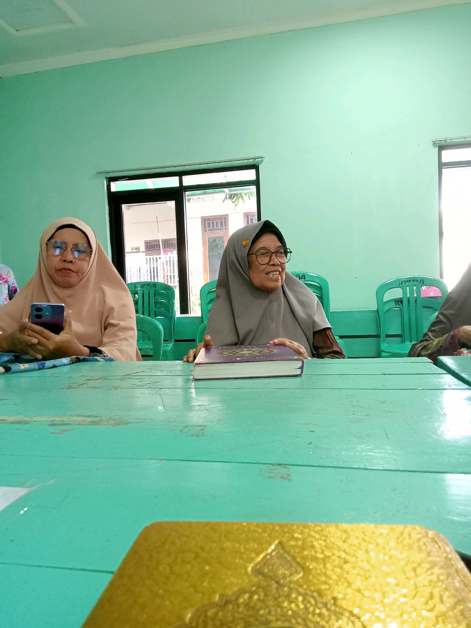 Two women wearing hijabs are seated at a long, turquoise wooden table in what appears to be a meeting room. The woman on the left wears a light brown hijab and glasses, and is holding a smartphone in her hands. The woman on the right wears a gray hijab and glasses, and is smiling and looking towards the left. She has a book with a purple cover in front of her. The room has pale green walls and a window with a view of a building with a tiled roof. Several green plastic chairs are visible behind the women. The overall impression is of an informal gathering or meeting.