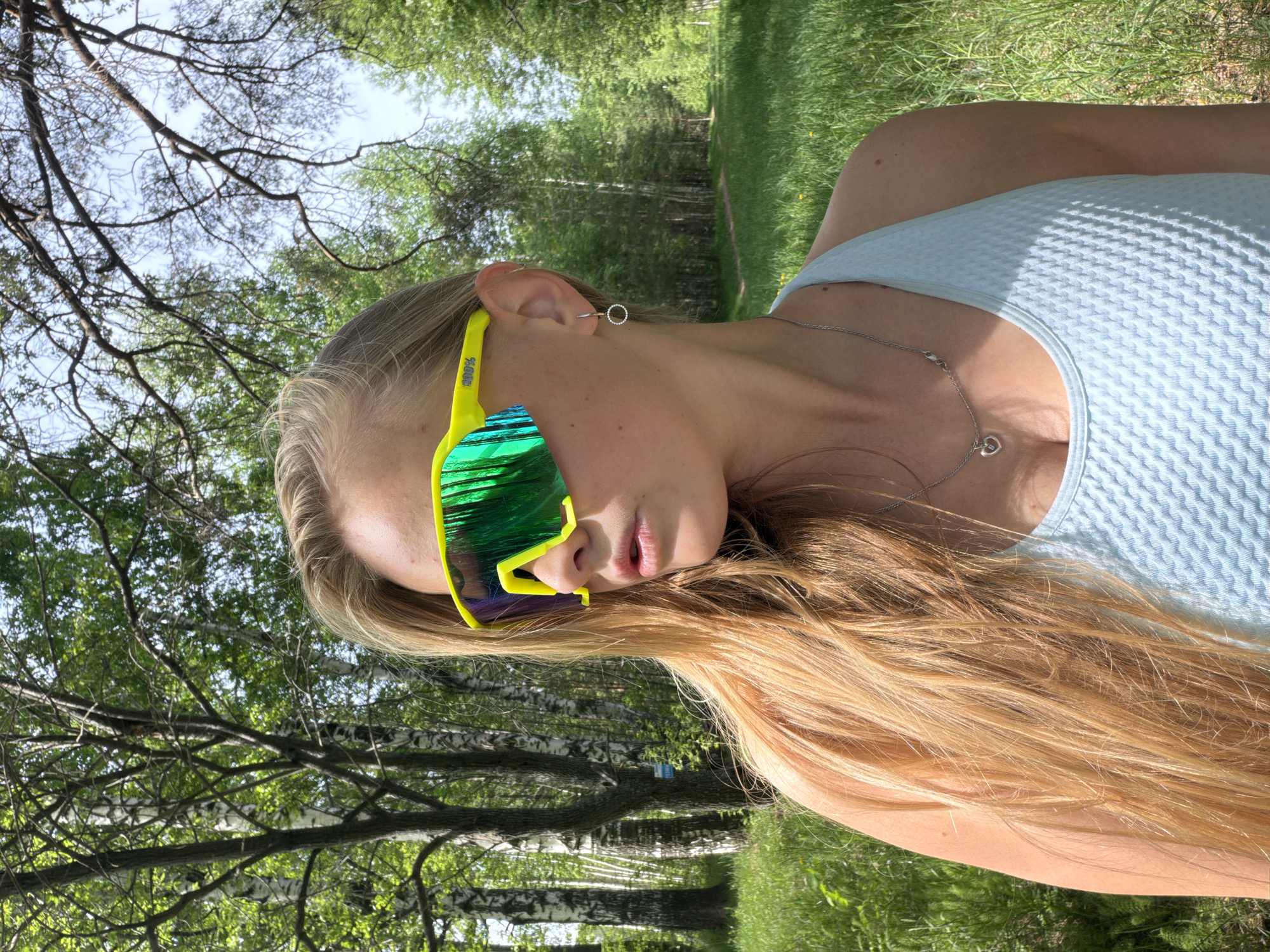 A young, fair-skinned woman with long blonde hair is facing the camera while wearing bright yellow sunglasses with green lenses. She is wearing a light blue textured top and a silver necklace with a heart pendant. Her ear is pierced in multiple places. The background shows a green, grassy area with birch trees and other leafy trees, suggesting a park or wooded area. The image appears to be taken outdoors on a sunny day. It is not possible to pinpoint the location to the city of Chaikovsky, Russia without additional information.