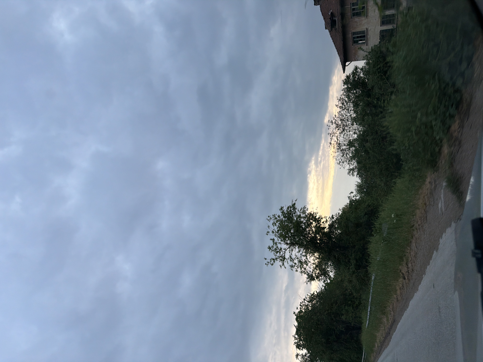 The image shows a rural roadside view. A paved road runs along the bottom right of the frame. Vegetation, including grass and trees, grows along the side of the road. In the background, a dilapidated building can be seen among the trees. The sky is overcast with a band of light appearing on the horizon. The location may be near the city of Oresh, Bulgaria, but there is no way to determine that definitively from the visual information alone.