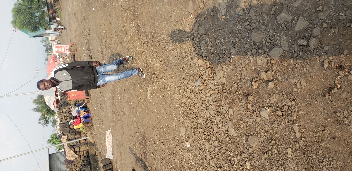 The image depicts an outdoor scene on an unpaved, dirt road in Buzove, Democratic Republic of the Congo. The ground consists of packed earth, gravel, and scattered rocks. In the mid-ground, a male, approximately 20-30 years old, walks forward towards the viewer. He is dressed in a black and grey athletic jacket with white stripes on the sleeves, faded blue jeans, and brown athletic shoes. His shadow is cast prominently on the ground towards the lower left of the frame. To the left, behind a low wall constructed from dark, irregular blocks, three individuals are seated on small chairs. One person is wearing a red top, another a yellow top. Adjacent to this wall, on the ground, several dark, rectangular blocks are stacked, and a light-colored patterned fabric lies flat. In the background, a structure with a green roof and a cross is visible to the right. Various other rudimentary buildings with corrugated metal roofs are present, along with sparse trees. Overhead, power lines or communication wires span the scene. Distant figures and a motorcycle are vaguely discernible further down the road. The scene is illuminated by direct sunlight, casting clear shadows.