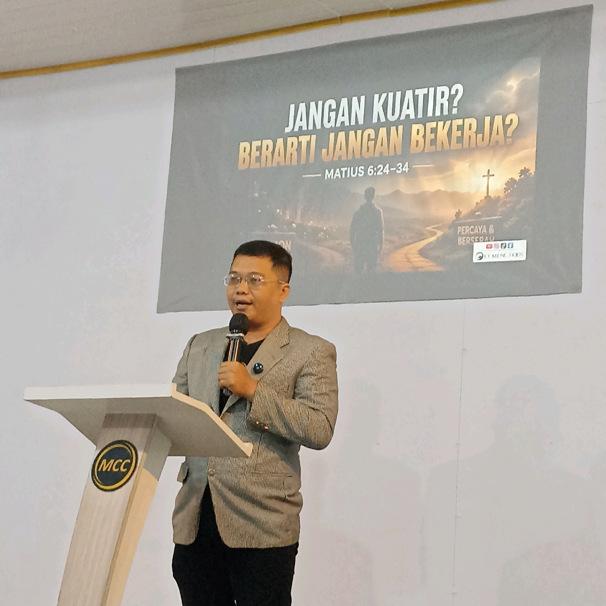 A man wearing a gray striped blazer, black shirt, and glasses stands behind a white podium and speaks into a microphone. The podium has a circular logo with "MCC" written in yellow on a black background. Behind the man, a large banner displays text in Indonesian that translates to "Don't worry? Means don't work?" and references Matthew 6:24-34. The banner also shows a silhouette of a person with a cross in the background. The scene appears to be indoors, possibly at a church or event hall, with a white wall and ceiling visible. The lighting suggests an indoor setting with artificial light. The man is the central subject, actively engaged in speaking.