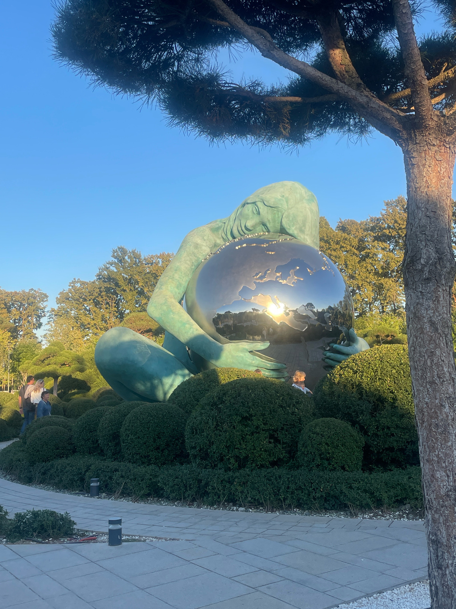 The image depicts a large, turquoise-colored sculpture of a reclining female figure cradling a shiny, spherical object that reflects the sun and the surrounding environment. The sculpture is situated in a garden with meticulously trimmed green bushes. In the background, several trees, including a large pine tree in the foreground on the right, contribute to the natural setting. The sky is a clear blue.
Several people are visible in the image. Two individuals, appearing to be adults, are standing on a paved walkway to the left of the sculpture. A child is partially visible amongst the bushes in front of the sculpture, looking up at it.
The scene appears to be outdoors, in a park or garden setting, possibly a public art installation. The style of the sculpture and the manicured landscaping suggest a place of aesthetic significance.
While the exact location in Krasnodar, Russia is not explicitly stated, the sculpture strongly resembles "The Motherland Calls" by sculptor Zurab Tsereteli, which is located in the Monument to the Defenders of the Fatherland in Krasnodar. This monumental work often depicts a female figure symbolizing the Motherland holding a sphere, which can be interpreted as the Earth or a symbol of peace and fertility.