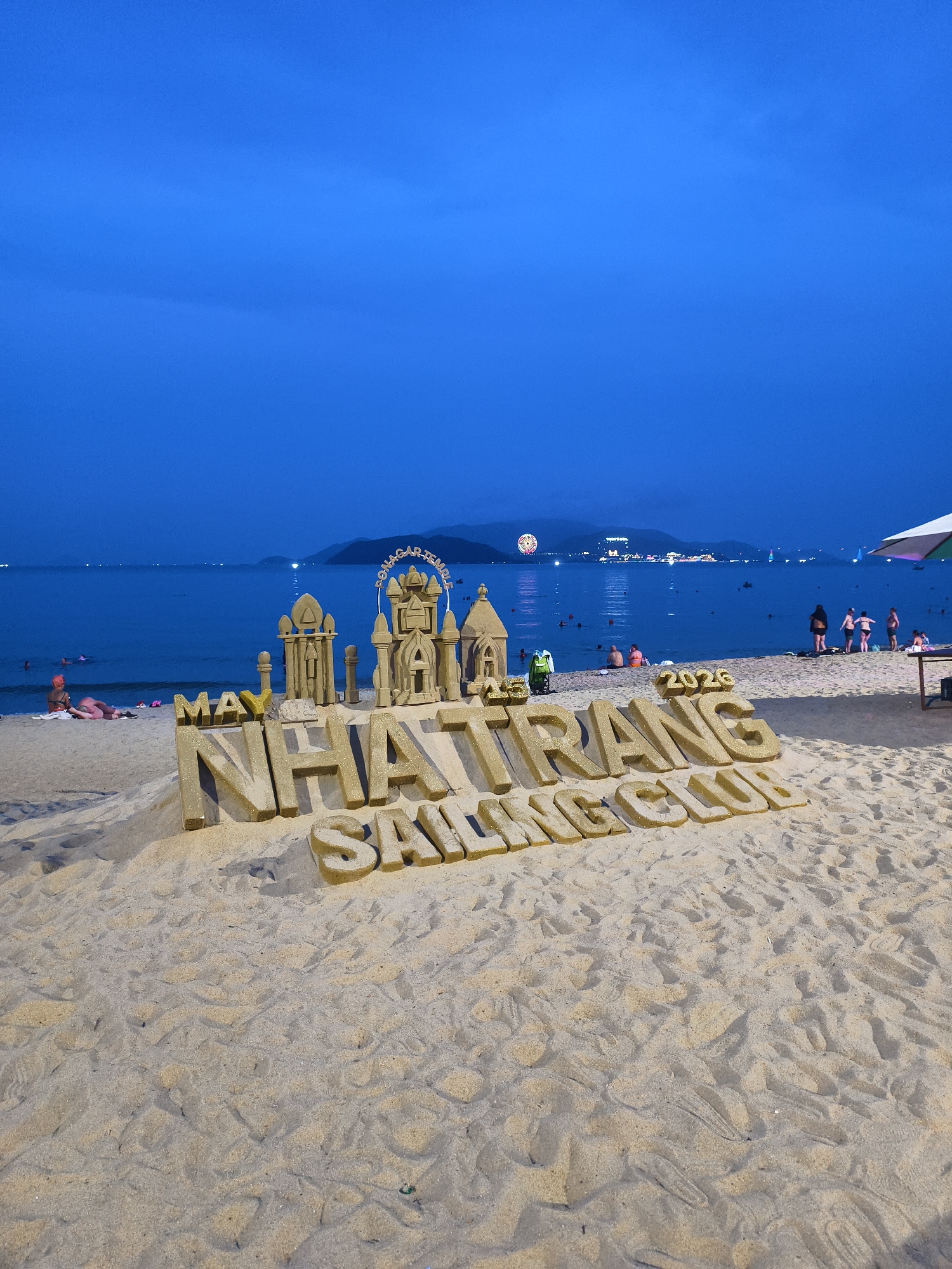 The image depicts a beach scene in Nha Trang Ward, Vietnam at dusk. A large sand sculpture is in the foreground, spelling out "MAY NHA TRANG SAILING CLUB 2026" with smaller sand structures resembling buildings or towers positioned behind it. Several people are visible in the background along the shoreline and in the water. A ferris wheel is visible in the distance, illuminated against the night sky. The overall scene suggests a festive or event-related atmosphere.