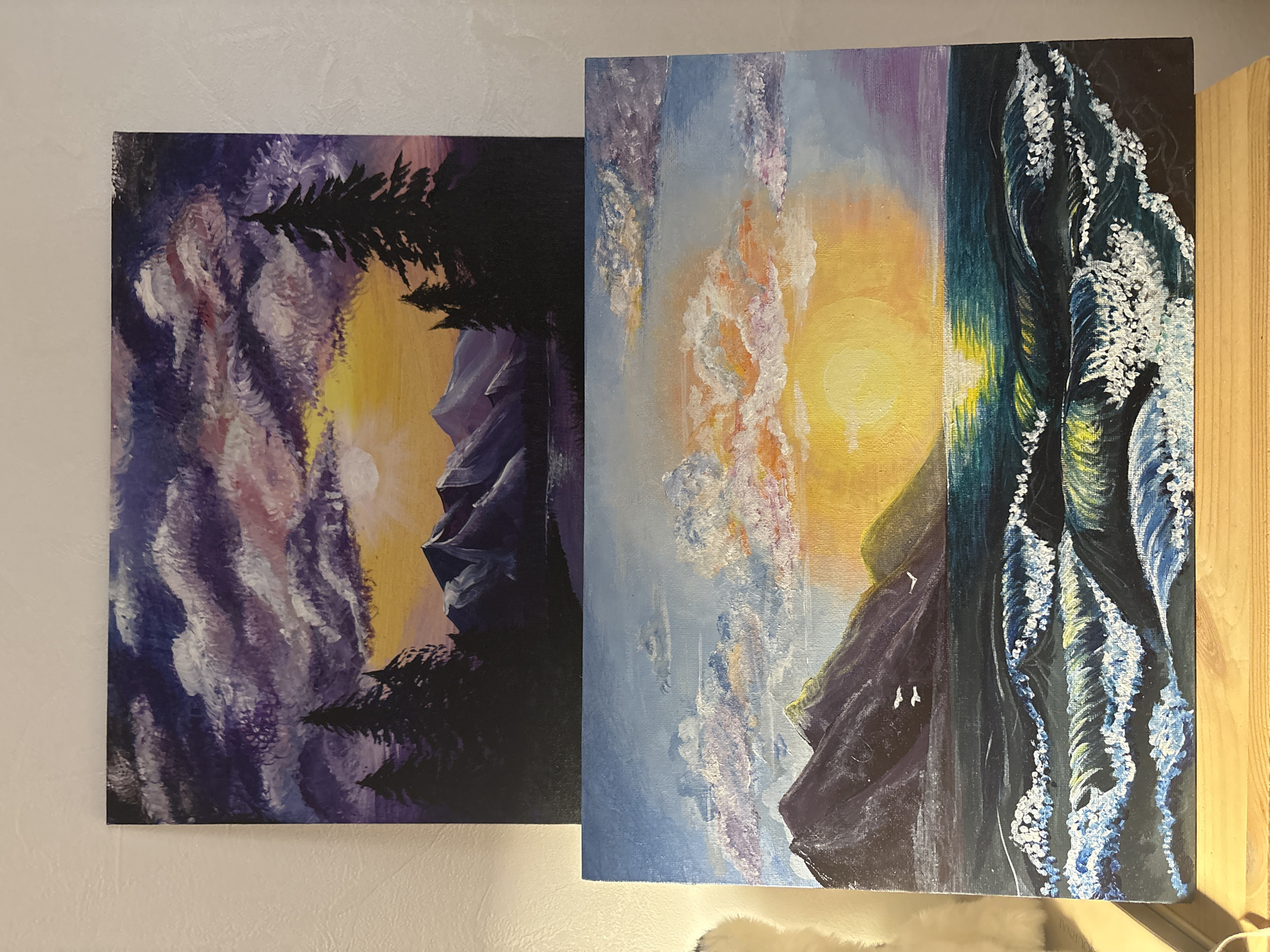 Two vibrant, stylized landscape paintings are leaning against a light-colored, textured wall. The images on the canvases appear rotated 90 degrees counter-clockwise from their natural upright orientation.
The painting on the left, which is slightly smaller, depicts a dramatic, atmospheric scene. The sky is filled with swirling clouds rendered in purples, whites, and soft pinks, with a central bright yellow and orange glow suggesting a sun or moon. Silhouettes of dark, coniferous trees frame the left and bottom right edges, while rugged, dark mountains are visible in the distance, bathed in a similar purple hue. The brushwork is expressive, giving a sense of movement to the clouds.
The larger painting on the right presents a dynamic seascape or coastal landscape. The upper portion shows a sky with a mix of light blue, soft purple, and sunlit orange and white clouds. A prominent yellow sun is positioned centrally, casting a bright glow. Below the sky, dark, rocky mountains or cliffs are depicted, with subtle green and yellow highlights. Towards the right side of the painting, a powerful ocean scene unfolds, featuring dark blue-green waves cresting with abundant white foam, conveying motion and energy. Two small white shapes, possibly birds, are seen flying near the mountains. The overall color palette of this painting is bright and contrasting, emphasizing the sunlit clouds and powerful waves against the dark landforms.
No people are visible in the image. No discernible text is present on the paintings or the wall. The wall texture is rough, and the scene is illuminated, possibly by natural light from the right, which creates a slight sheen on the surface of the paintings.