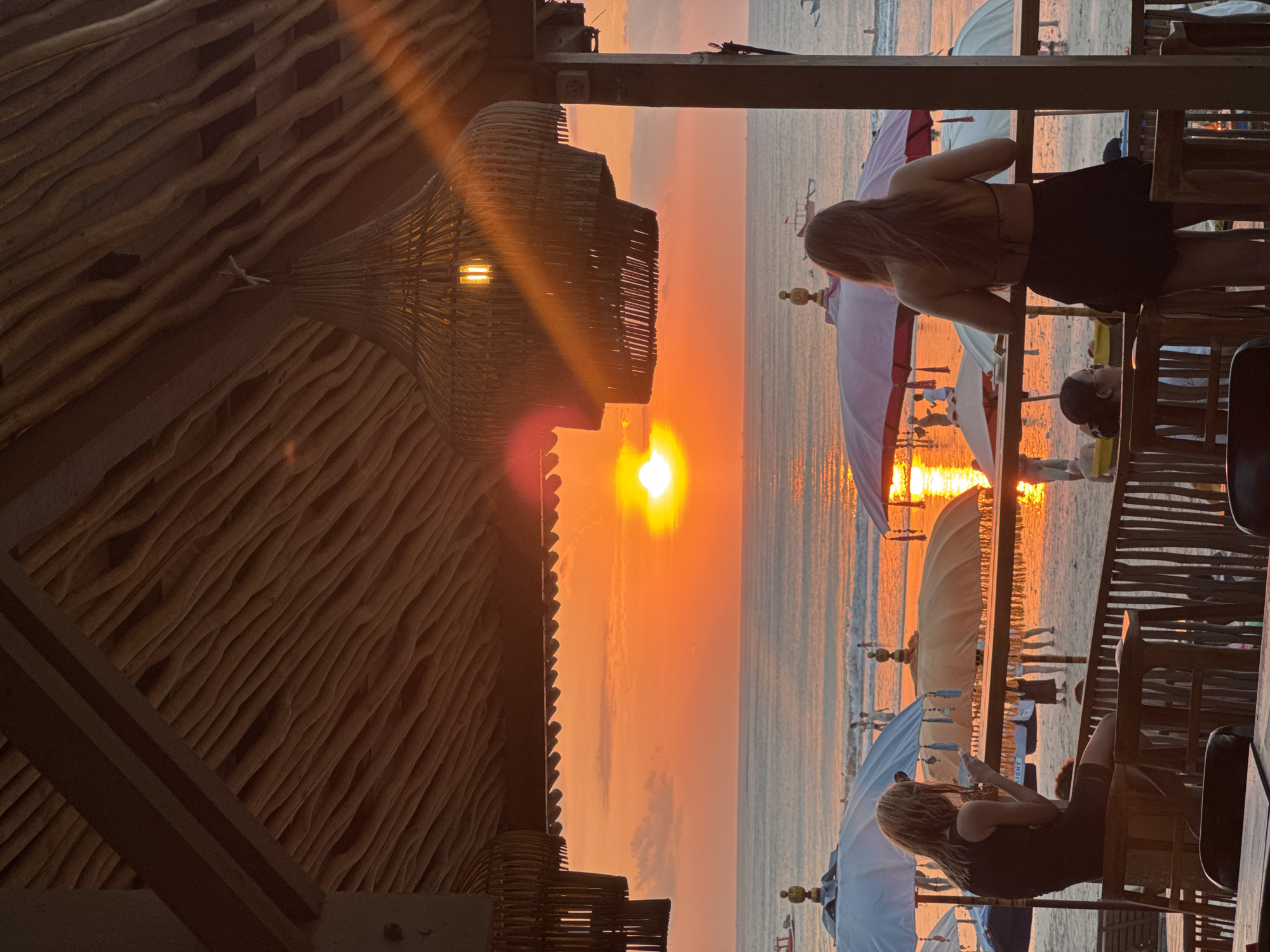 The image captures a picturesque sunset scene from within a rustic, wooden beachfront establishment in Kuta Selatan, Indonesia. The golden-orange sun, a prominent focal point, is positioned low on the horizon, casting a brilliant, warm glow across the sky and reflecting intensely on the calm ocean waters and the wet sand of the beach.
In the foreground, two individuals, both women, are viewed from behind, engrossed in the spectacle of the setting sun. On the left, a woman with long, light-colored hair stands, leaning contemplatively on a sturdy wooden railing, looking out towards the horizon. To her right, another woman with long, dark hair is seated at a wooden table, also facing the sunset, possibly interacting with a mobile phone.
The interior of the structure, occupying the left side of the frame, features a distinctive textured wooden ceiling or wall made from natural, undulating wooden planks or logs. A large, intricately woven lamp fixture hangs down, emitting a warm, artificial light that harmonizes with the natural light of the sunset. Wooden beams and other structural elements complete the cozy, natural aesthetic.
Beyond the railing, the beach extends, adorned with several traditional-style beach umbrellas—some white, others light pink or purple—which create beautiful reflections on the glistening, wet sand. Several other indistinct figures of people can be seen enjoying the beach in the distance. The scene evokes a sense of tranquility and natural beauty, characteristic of a tropical sunset experience. A strong lens flare emanating from the sun adds to the warm, ethereal atmosphere of the photograph. No visible text is present.