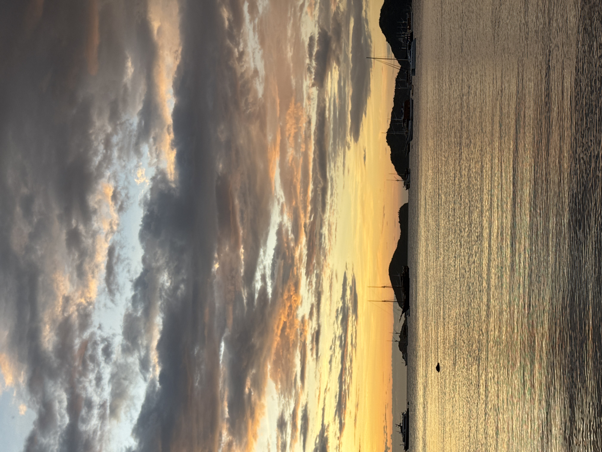 Captured at an unknown location in an unknown city, the scene portrays a seascape at sunset. The sky is dominated by heavy, layered clouds ranging from dark grey to light white, tinted with golden hues from the setting sun. The horizon line is visible, with silhouettes of land and boats against the yellow sky. The water reflects the golden light, creating a shimmering effect. No people are visible.