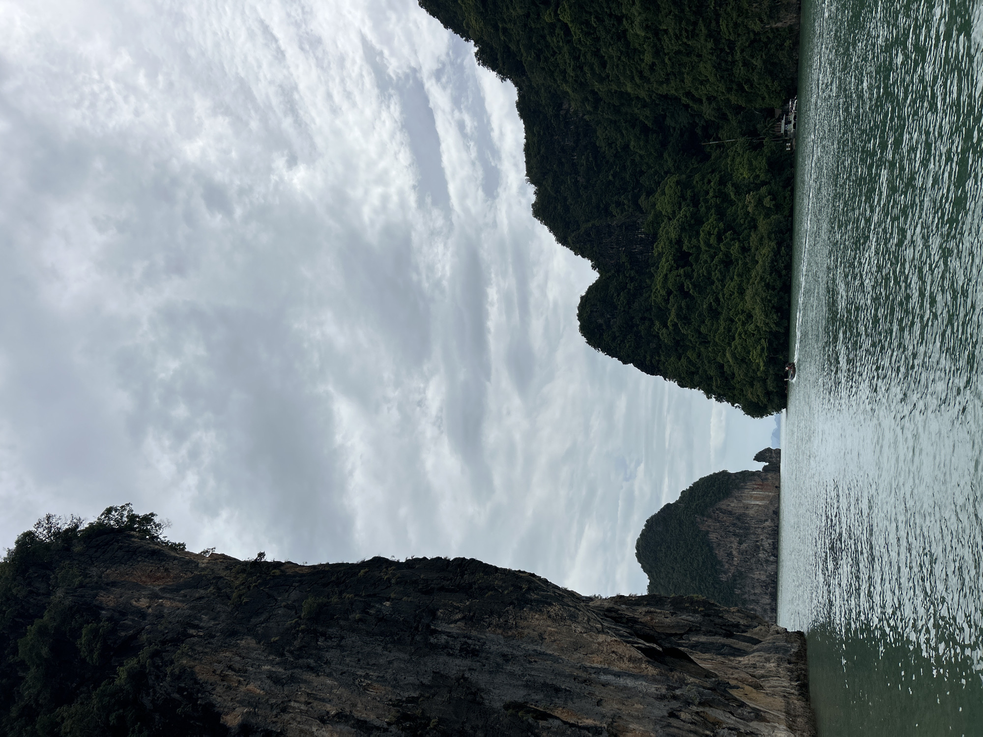This image depicts a scenic view of a body of water surrounded by lush green islands and towering cliffs. The water appears calm with gentle ripples, reflecting the overcast sky. In the distance, several small boats are visible on the water, suggesting a recreational or travel context. The islands are densely vegetated, with trees covering their steep slopes. The cliffs on the left side of the image are rugged and rocky, with some sparse vegetation growing on them. The sky is predominantly cloudy, with patches of lighter clouds suggesting diffused daylight. The scene captures a natural landscape, likely a tropical or subtropical coastal area. While the image has been described as being from Krasom, Thailand, this specific location is not visually identifiable from the image alone.