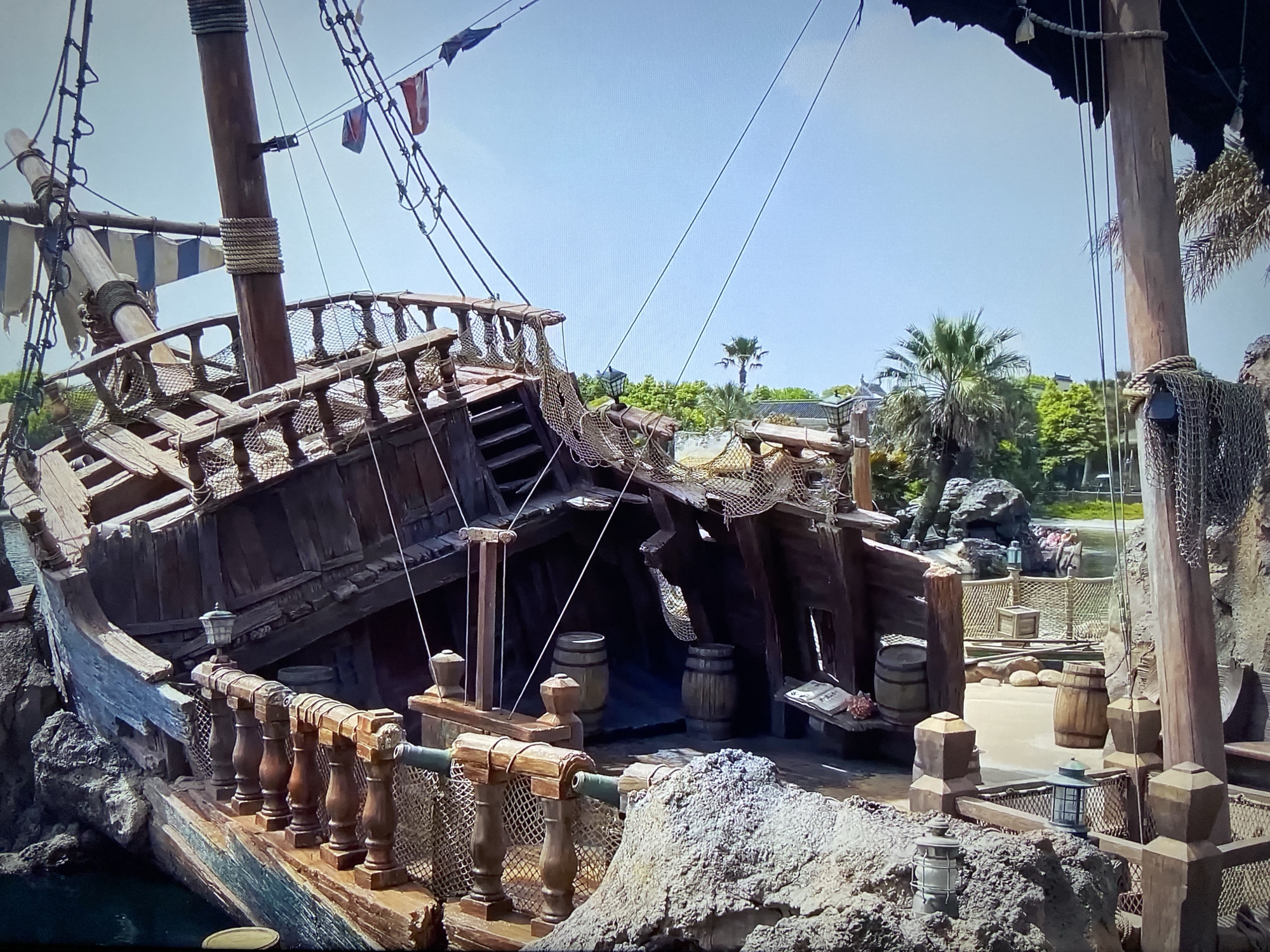 The image shows a section of a shipwrecked wooden pirate ship in what appears to be a themed area of a park. The ship is tilted and appears heavily damaged, with broken planks and rigging. There are barrels visible on deck. In the background, there are palm trees and other greenery, suggesting a tropical or subtropical environment. The scene appears to be located in Tokyo DisneySea, Tokyo, Japan, specifically in the area known as "Pirate's Shores". The photo looks like it was taken from on land or a dock.