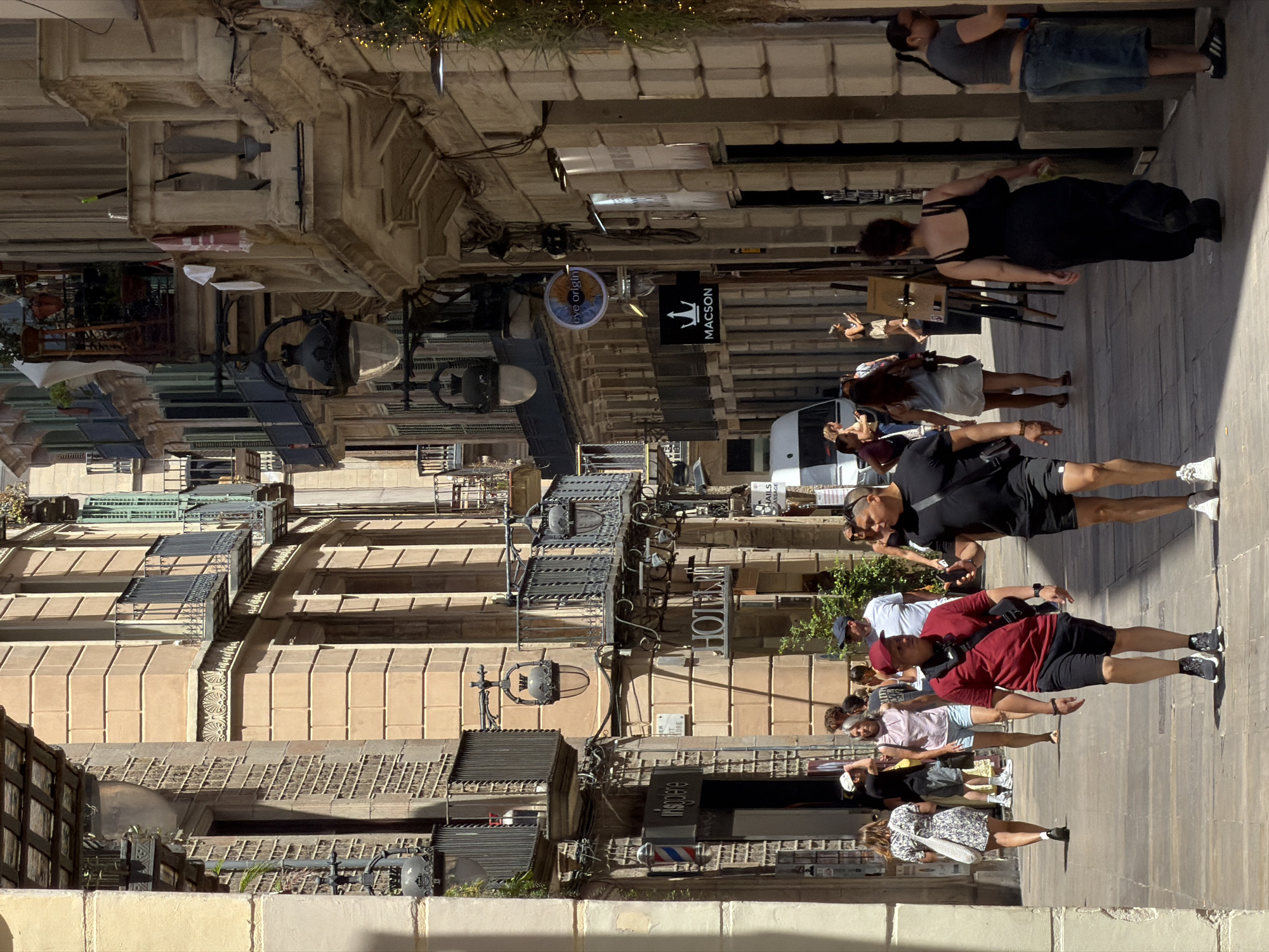 This media file shows a street scene in Barcelona, Spain. The orientation of the image is landscape and it appears to be taken during the day, under bright sunlight, suggesting fair weather.
The primary subjects are several individuals walking or standing on a paved street lined with buildings. The architecture is detailed, with stone facades, balconies, and ornate windows, suggesting an older, possibly historic part of the city. Some buildings have businesses with signage. One sign reads "HOTEL PASEO," another "MACSON," and there's a circular sign with the words "eve origin" and a sun-like graphic. There's also a barber pole.
The people are dressed in casual summer attire. Some are looking at their phones, while others appear to be conversing or looking around the street. One person is wearing a black dress, another a red shirt and shorts, and several others are in t-shirts and shorts or skirts. The overall mood seems relaxed, typical of a leisurely stroll through a city.
In the background, there are glimpses of more buildings and possibly some greenery. The scene is populated but not overly crowded, allowing for clear views of the individuals and the surrounding architecture. The quality of the image is good, with sharp details and accurate colors, despite the unusual vertical orientation.