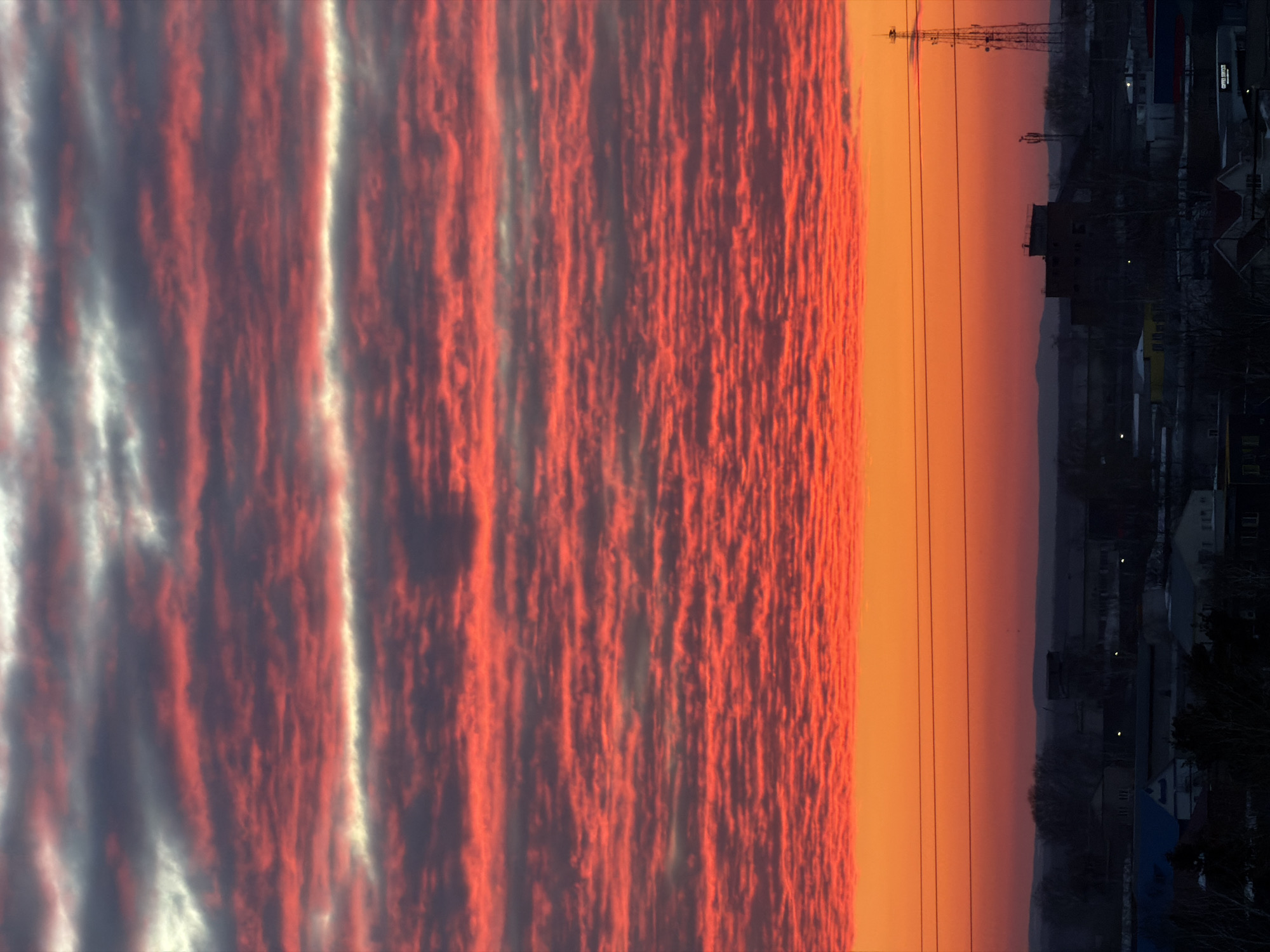 The image displays an urban landscape at either sunrise or sunset over Chaikovsky, Russia. The sky dominates the upper two-thirds of the frame, characterized by horizontal bands of altocumulus clouds. These clouds are predominantly saturated with bright red and orange hues, interspersed with darker grey segments and narrow, horizontal streaks of white and light grey. The intensity of red and orange fades towards the top of the frame. Below the cloud layer, the sky transitions from a vibrant orange at the cloud base to a darker orange and deep purple-red near the horizon.
In the lower third of the image, the city skyline is silhouetted against the colorful sky. The foreground depicts a mix of dark, indistinct residential and industrial buildings. Several power lines or utility cables stretch horizontally across the mid-ground, casting thin, dark lines against the bright orange sky. On the far right, a tall, lattice-structure telecommunications tower is prominently visible. To its left, a multi-story, dark industrial or commercial building stands out. Scattered points of artificial light, likely from buildings, are visible within the dark urban structures. Distant hills or elevated terrain are faintly discernible as a dark silhouette on the far horizon. No people, specific actions, or interactions are clearly visible.