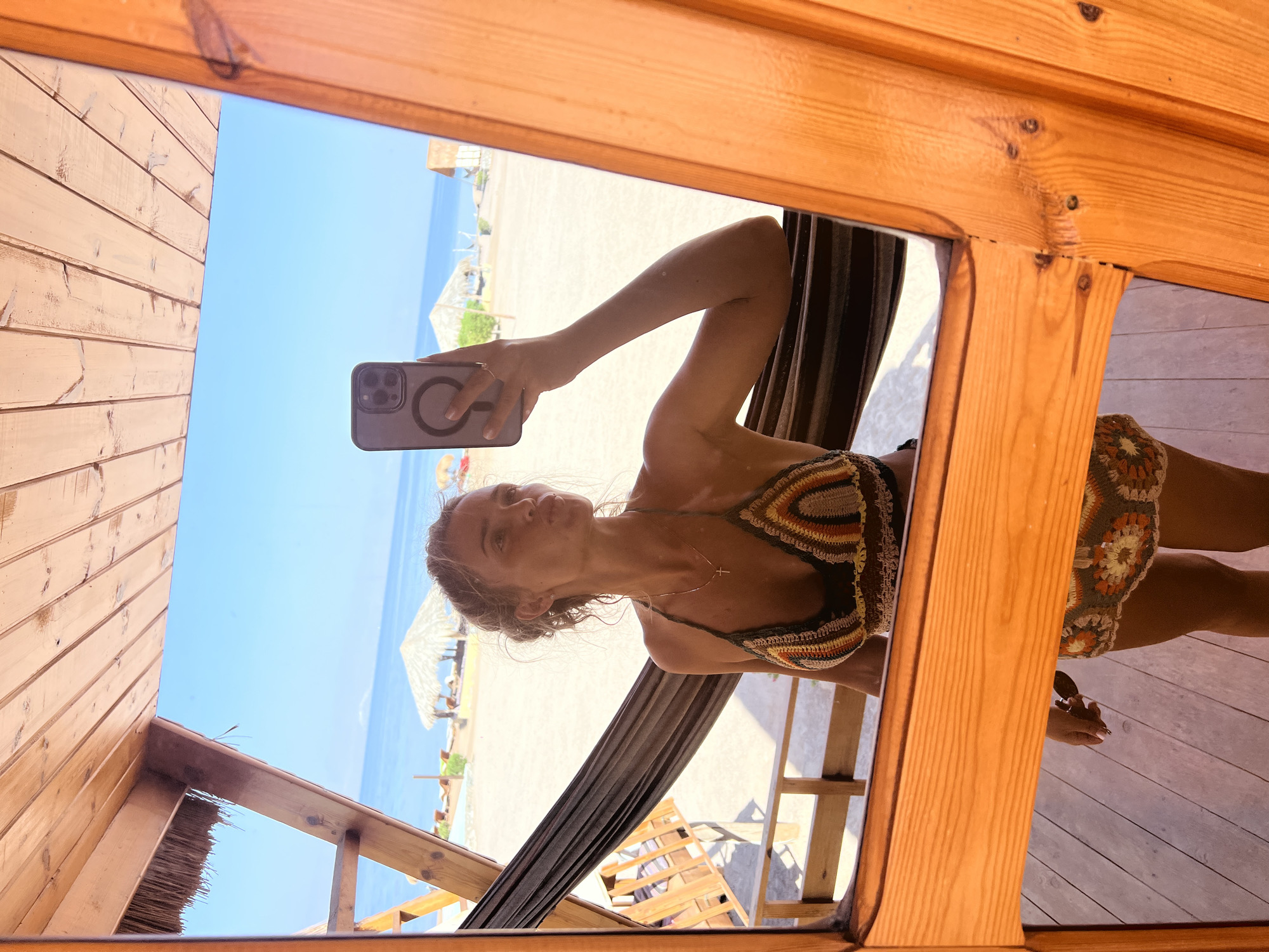 A young woman with blonde hair pulled up into a bun and wearing a crocheted bikini and skirt is reflected in a wooden-framed mirror. She is holding a phone in her right hand and has her left hand resting on a hammock. The background of the reflection shows a beach with palm trees, umbrellas, lounge chairs, and the sea under a clear blue sky. The foreground shows the wooden structure of the room the mirror is in, with visible wooden floorboards. The scene is likely in Nuweiba, Egypt, given the beach setting and typical resort architecture.
