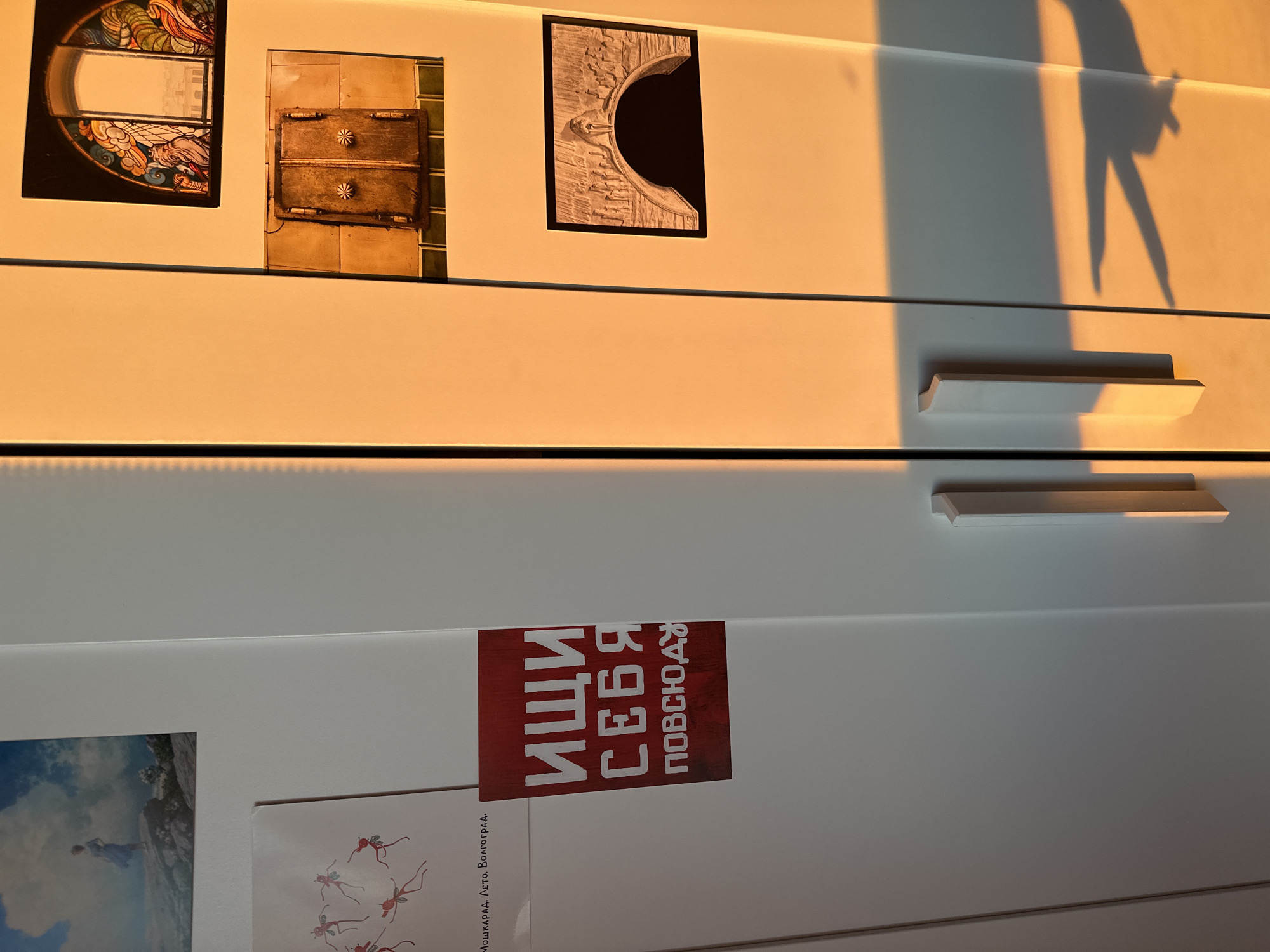 The image shows the side of a white cabinet, possibly a refrigerator, with several items attached to its surface. The cabinet has two handles and is illuminated by warm, golden light, casting long shadows across it. On the left side of the cabinet, there is a postcard with a painting of a woman standing on a rocky outcrop against a cloudy sky. Below that, another postcard depicts a circular arrangement of small, dancing figures. Further down, a red sticker with white Russian text reads "ИЩИ СЕБЯ ПОВСЮДУ," which translates to "Seek yourself everywhere." On the right side of the cabinet, three more items are attached. The top one is a rectangular image of a stained-glass window with an arched top, featuring colorful figures. Below it, a square image shows a weathered, metallic double door with decorative circular handles. The bottom image on the right is a rectangular illustration of an arched structure, possibly an aqueduct or bridge, with a procession of figures. The overall scene suggests an indoor setting with natural light, and the presence of multiple decorative items indicates personal belongings. The image does not contain enough information to determine its specific location within Moscow, Russia.
