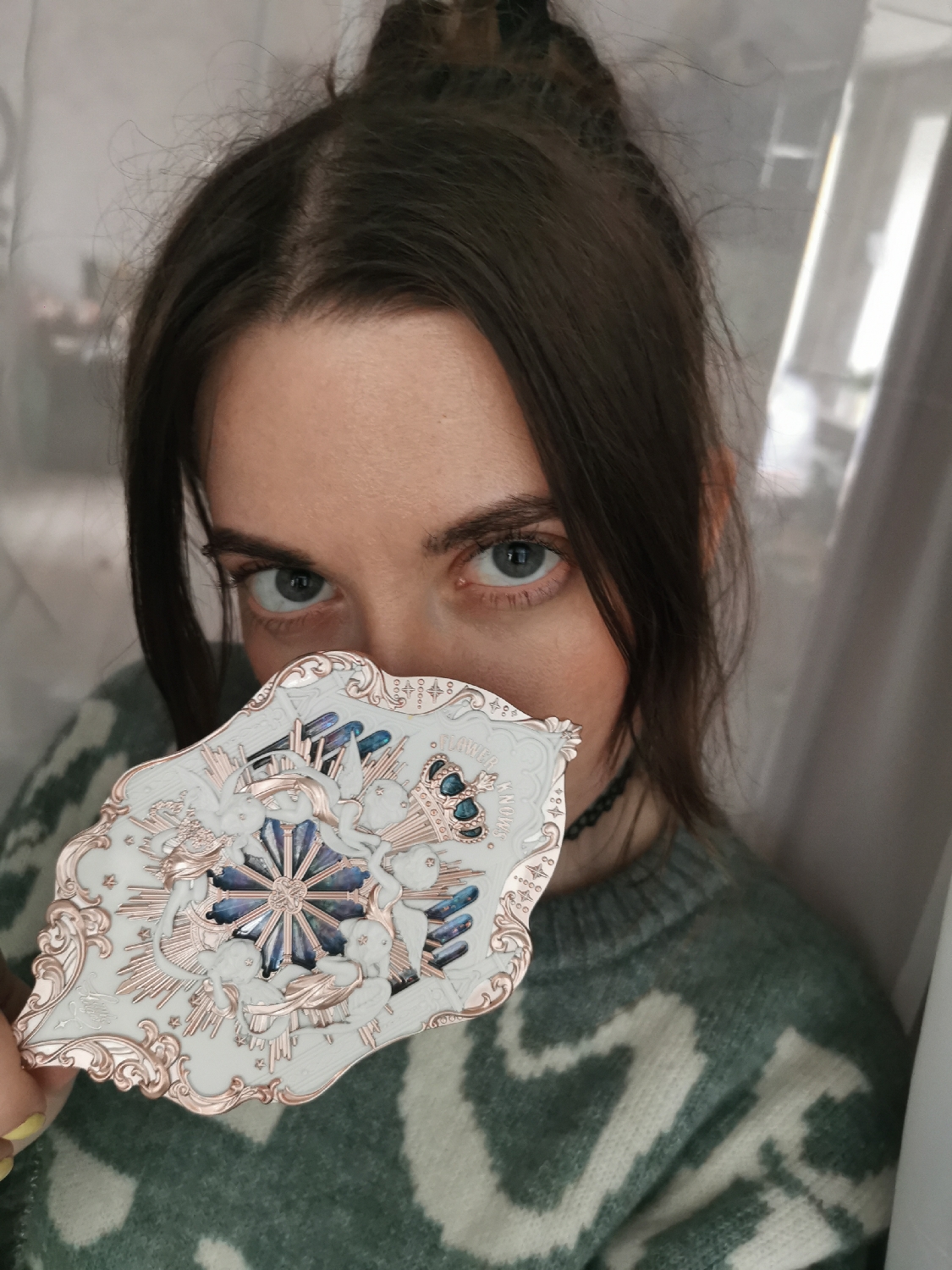 A young woman with dark brown hair, styled in a messy top bun with loose strands framing her face, looks directly at the camera. Her blue-green eyes are prominent, while the lower part of her face is obscured by an ornate, decorative object she is holding. She has fair skin and visible eyebrows.
She is wearing a cozy-looking sweater with a two-tone pattern of green and off-white, possibly a marled or patterned knit. Around her neck, a thin, dark choker or necklace is visible. The object she holds is a highly detailed, white and rose-gold colored compact or handheld mirror with an irregular, rococo-inspired shape. Its design features cherubic figures, a prominent crown, and a central radiating star-like motif with blue and iridescent facets. Faint text reading "FLOWER" and possibly "ANONIA" is visible on the object's surface, along with small, stylized "OOO" patterns.
The background is indistinct, appearing to be an indoor setting with soft, light-colored fabric or curtains, creating a simple and uncluttered environment. The lighting is soft and even, highlighting the details of the object and the woman's eyes. The overall activity involves the woman presenting this decorative item while maintaining eye contact with the viewer.