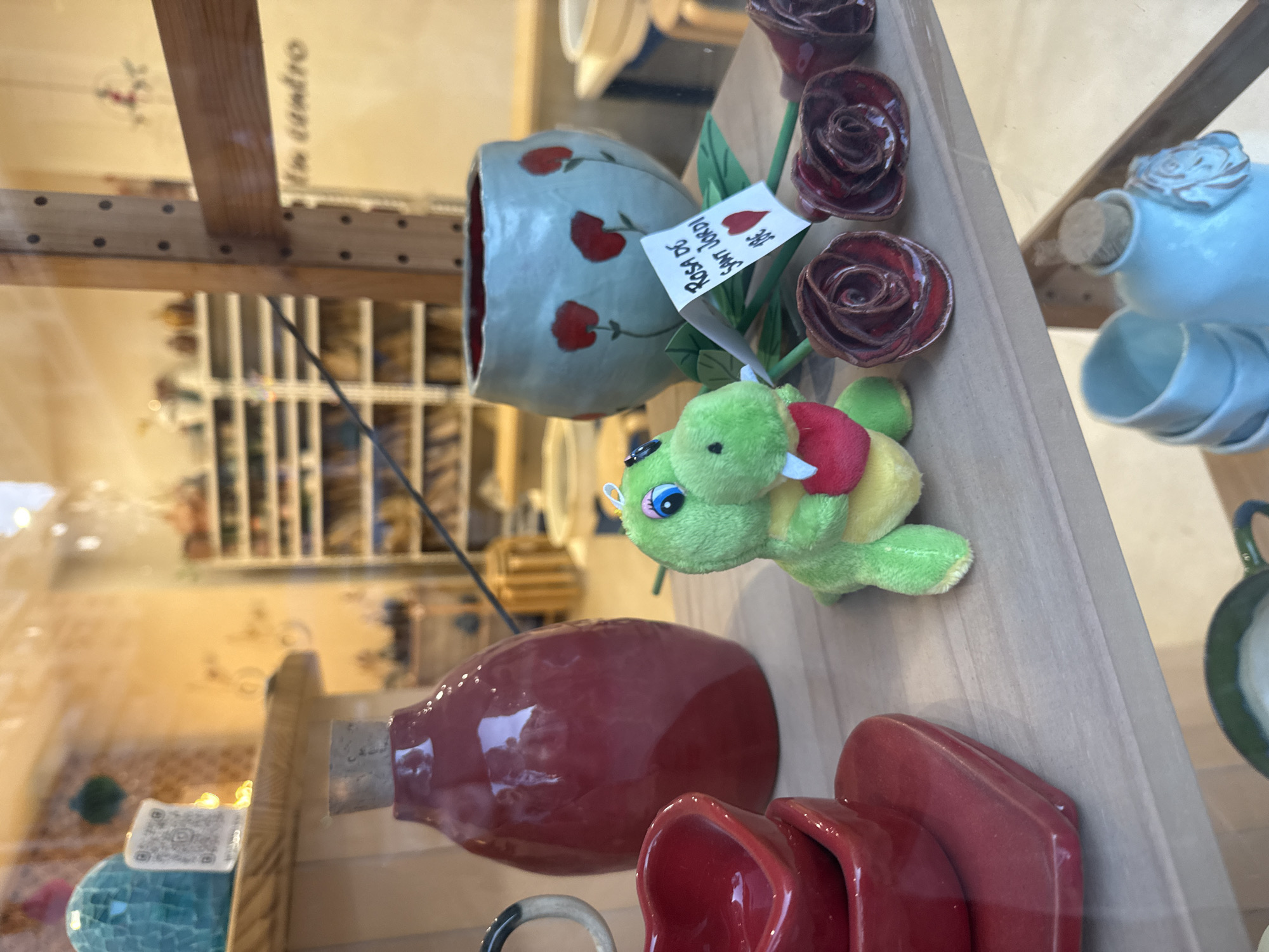 This image, captured in Barcelona, Spain, presents a tilted view of a retail display featuring ceramic items and a plush toy on a light wooden shelf, likely seen through a shop window. The scene is brightly lit, with visible reflections on the glass.
Central to the display is a light blue-grey ceramic vase adorned with red hearts. A white tag attached to it clearly reads "ROSA DE SANT JORDI" (Rose of Saint George) and "10€", indicating its price and relevance to Saint George's Day, a significant cultural event in Catalonia. Adjacent to the vase are several dark red ceramic roses with green stems. A small green plush toy, resembling a turtle or dragon and featuring a red heart on its back, is positioned alongside the roses.
Further to the right, a light blue ceramic bottle or pitcher, topped with a rose-shaped stopper, is visible, accompanied by a smaller light blue ceramic cup or bowl. On the lower part of the shelf, a glossy, dark red ceramic bottle or vase is displayed, with several deep red ceramic cups or bowls stacked beneath it. A spherical object with a blue mosaic pattern and a small QR code sticker are also partially visible on the bottom left.
The background is softly blurred, revealing additional shelves and merchandise, suggesting the interior of a pottery or souvenir shop. The overall presentation is a curated display of handcrafted items, clearly themed for the Sant Jordi celebration, where roses are a traditional gift.