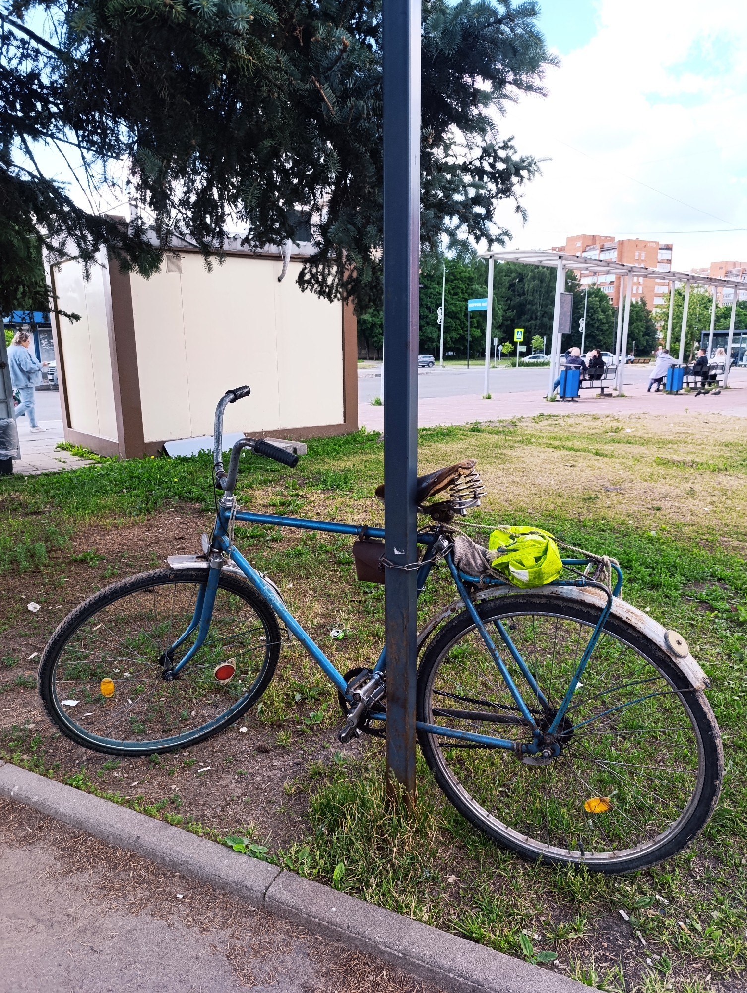 In an urban outdoor setting, a blue bicycle is chained to a dark grey metal pole. The bicycle has a brown leather seat with visible springs, a rear rack with a bright green bag and what appears to be some rolled-up documents or papers. The bicycle also has a brown saddlebag attached to the frame. The bicycle is parked on a grassy area next to a paved sidewalk. In the background, a beige building with brown trim is visible, along with a bus stop shelter where several people are seated. Further in the distance, brick buildings and trees are present, under a cloudy sky. A young woman in a light blue jacket and jeans walks past the beige building on the left side of the image.