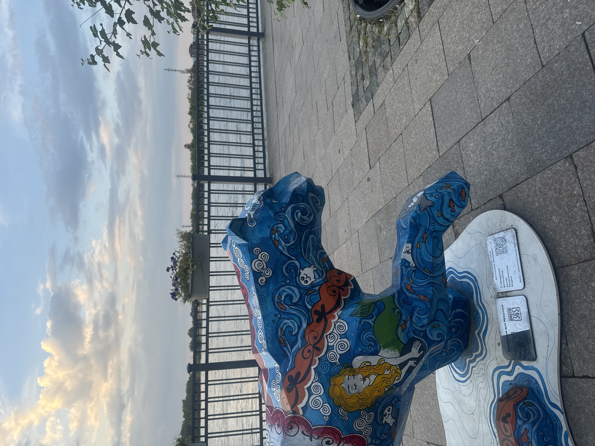 The image shows a decorated sculpture of a stylized animal, possibly a tiger or a panther, placed on a paved walkway next to a body of water. The sculpture is painted with a blue and orange theme, depicting waves, fish, clouds, and a mermaid with blonde hair. There are two information plaques and a QR code attached to the sculpture's base. The background reveals a metal fence, a planter with flowers, and a wide river under a cloudy sky. In the distance, trees and some buildings are visible, along with power lines. The overall scene suggests an outdoor public space, likely a waterfront promenade in the city of Irkutsk, Russia.