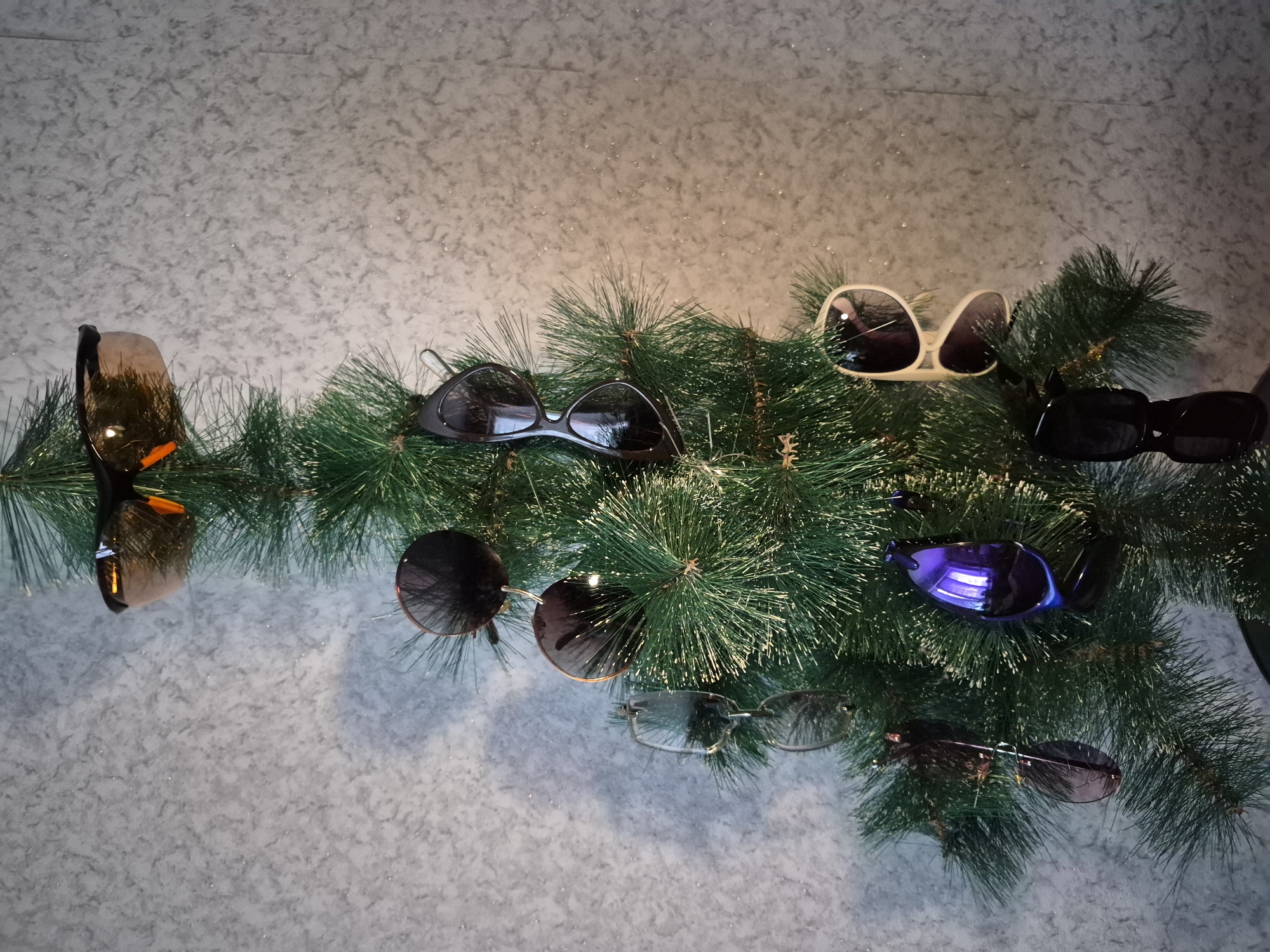A top-down shot depicts a variety of sunglasses arranged on an artificial evergreen garland, placed against a backdrop of mottled gray-white material. The sunglasses exhibit various styles and colors: orange-lensed sports glasses, classic black sunglasses, beige-rimmed sunglasses, round sunglasses with copper-tone rims, thin-rimmed glasses, and a pair with blue lenses. The setting and location are not determinable from the provided image.