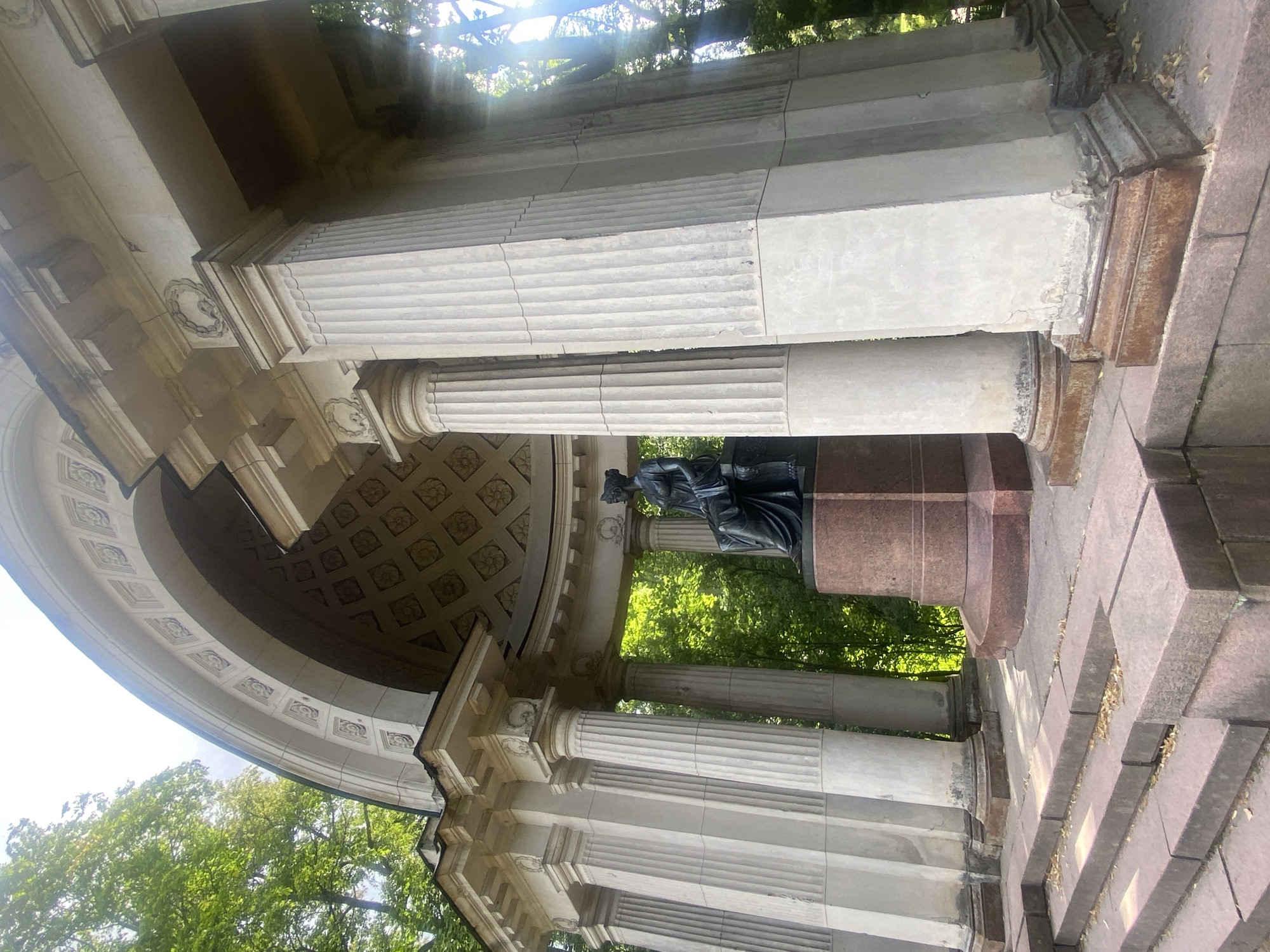 This is a low-angle, full shot of a classical-style rotunda structure within a park setting, likely in Pavlovsk, Russia, given its architectural style and prevalence of trees. The rotunda features a semi-circular archway supported by fluted Doric columns. The arch's interior is decorated with a grid of square panels, each containing a stylized floral motif. The exterior of the rotunda is adorned with decorative wreaths above the columns.
At the center, within the rotunda, sits a dark bronze statue of a seated woman dressed in classical attire. She is positioned on a two-tiered, reddish-brown pedestal. The ground in front of the rotunda consists of broad, tiered steps made of the same reddish-brown stone. The surrounding environment is lush with green foliage, suggesting a well-maintained park or garden. Sunlight filters through the trees, creating dappled light and lens flare effects in the upper right portion of the image. The stone of the rotunda shows signs of age and weathering, with some staining and chipping visible.
