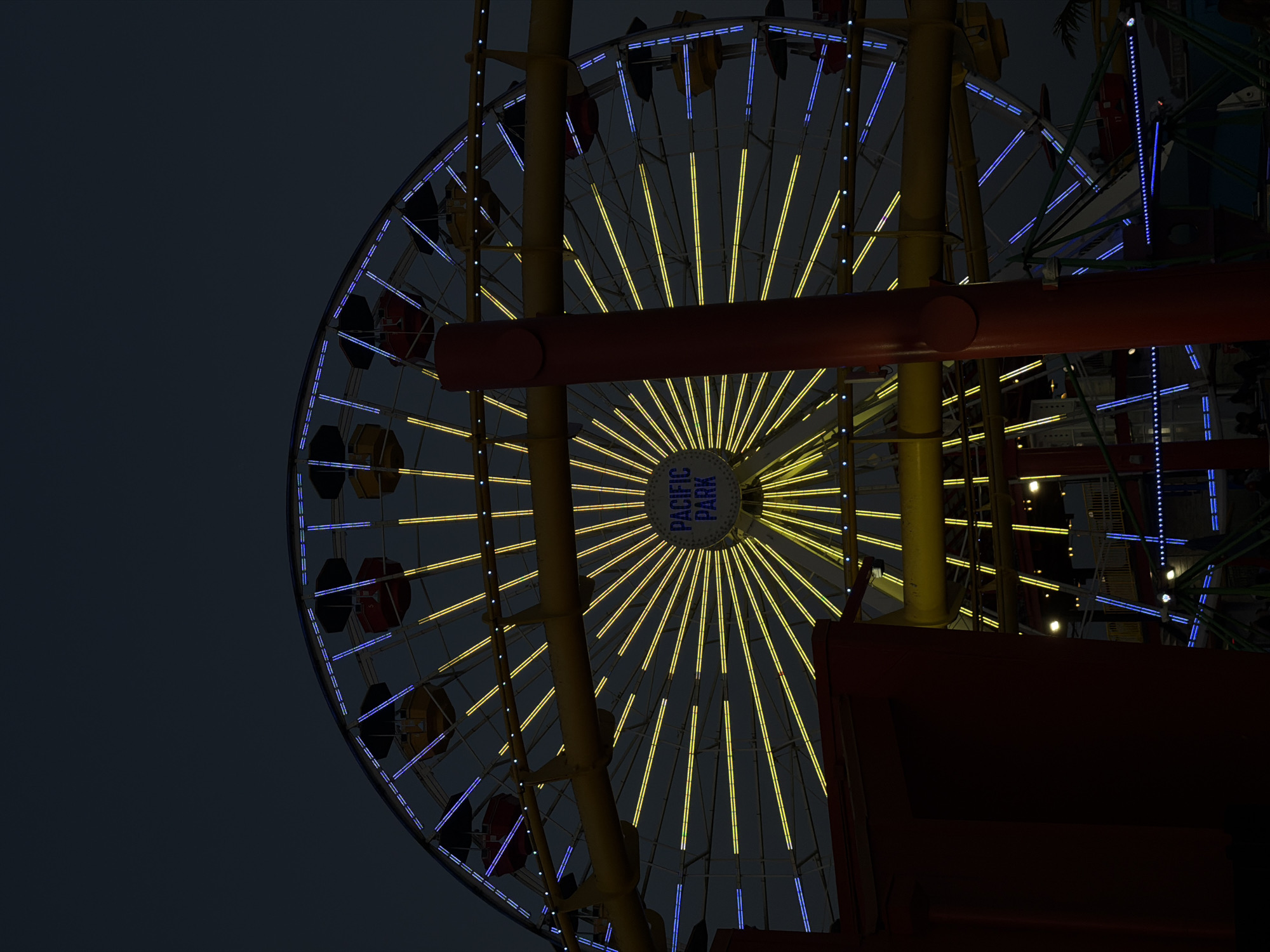The image captures a partial view of the illuminated Ferris wheel at Pacific Park on the Santa Monica Pier in Santa Monica, California, United States of America, at night. The wheel is adorned with yellow and blue lights. The lower portion of the image is partially obscured by the pier structure. The sky is a dark gray.