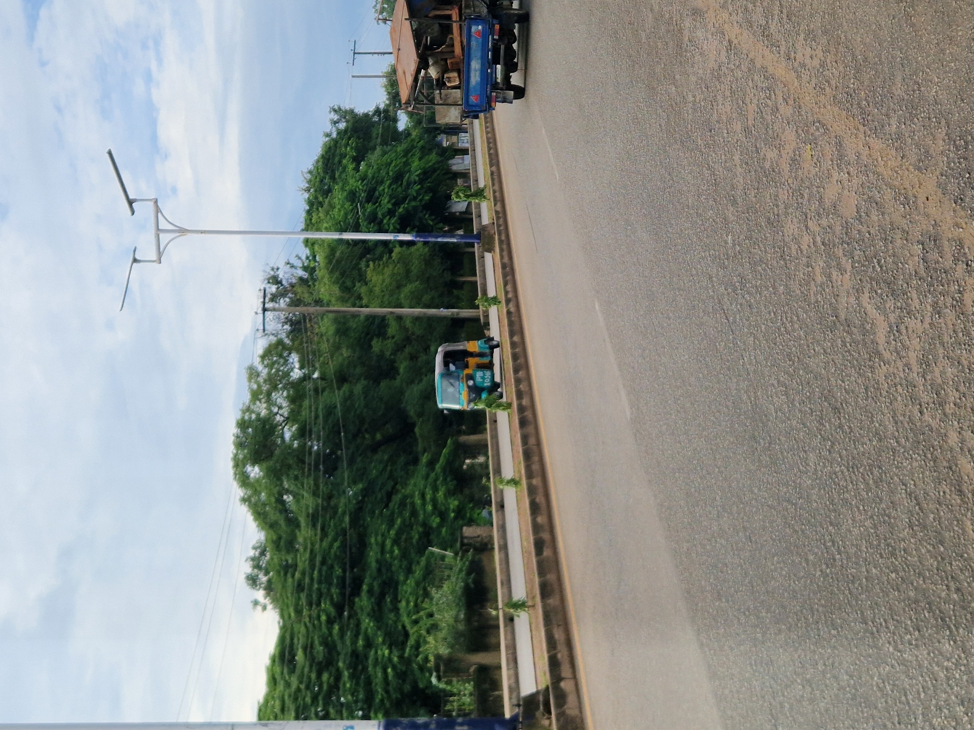 This image depicts a street scene in New Stadium, Nigeria. A two-lane paved road is visible, with a median strip separating lanes of traffic. On the left side of the road, a light pole with a solar panel is present, and beyond that, a dense line of trees forms a backdrop. In the middle distance, a teal and yellow auto-rickshaw is traveling on the road. To the right of the auto-rickshaw, a blue flatbed truck is also moving along the road, carrying lumber and other materials. The sky above is partly cloudy.