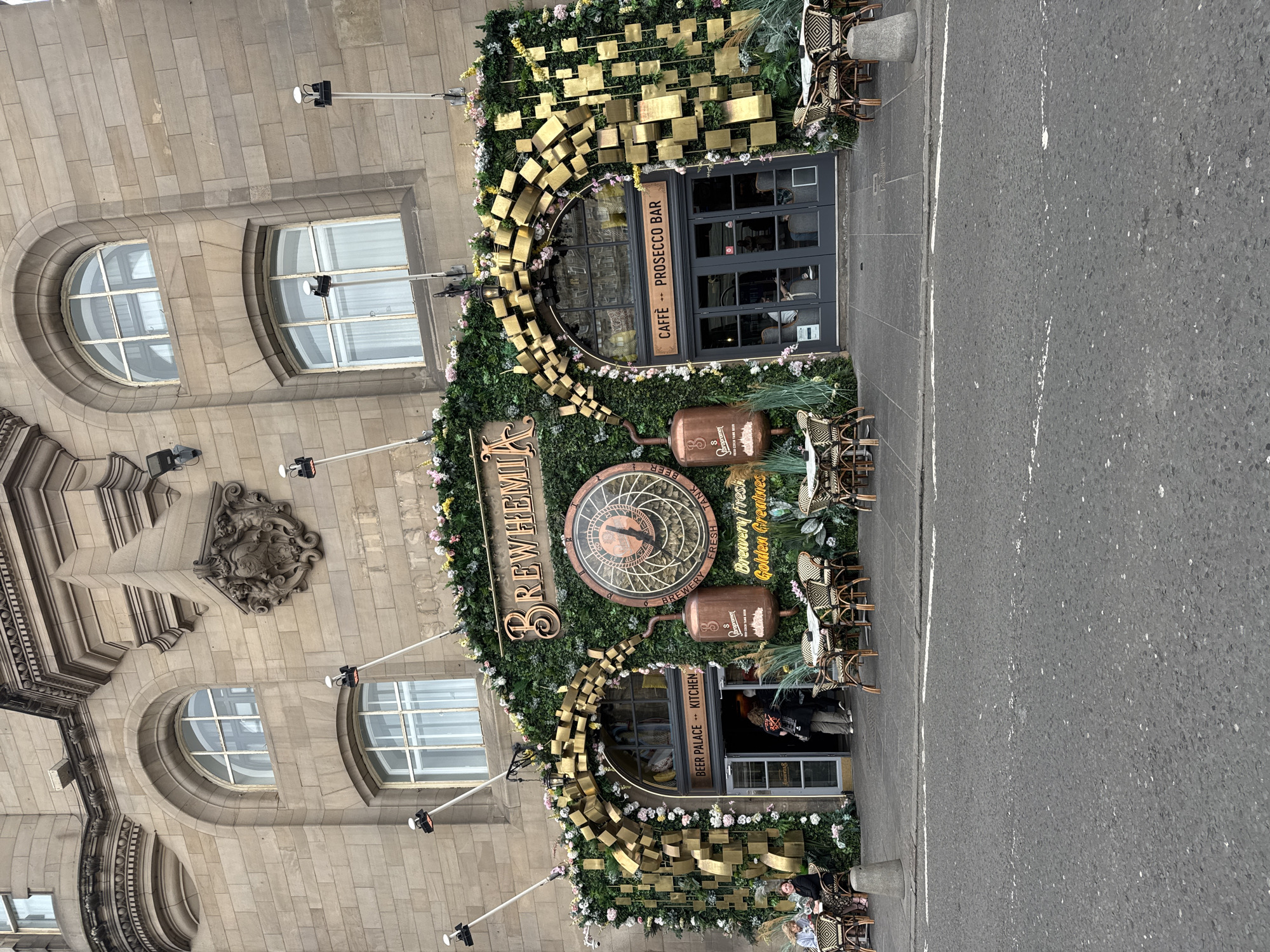 The image shows the exterior of a building, identified as "BREWHAMIA", located in the city of Edinburgh, United Kingdom. The building's facade is made of stone and features arched windows. The entrance is heavily decorated with artificial greenery, flowers, and golden geometric shapes. A large clock with a brewing-themed design is prominently displayed. Outdoor seating with tables and chairs is arranged in front of the entrance, with some patrons seated at the tables. The street in front of the establishment is paved, with white lines indicating traffic lanes. Several people are visible, including patrons seated at tables and individuals entering or exiting the building. The overall scene depicts a bustling outdoor dining and drinking establishment.