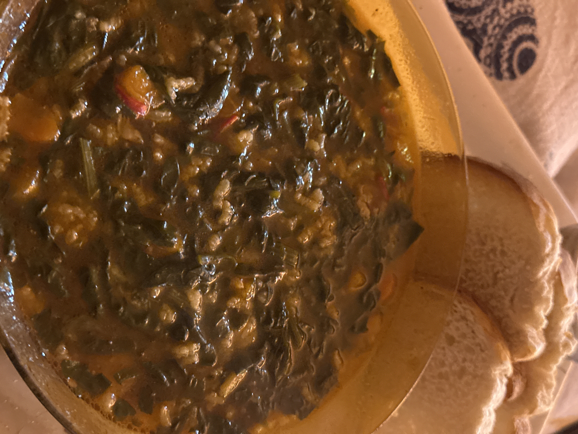 A high-angle shot shows a bowl of dark, greenish-brown soup alongside slices of white bread on a light-colored cloth. The soup appears to contain leafy greens and small pieces of vegetables. The bowl is made of yellow glass or plastic, showing visible surface imperfections. Part of the cloth has a simple, dark-blue and white patterned design.