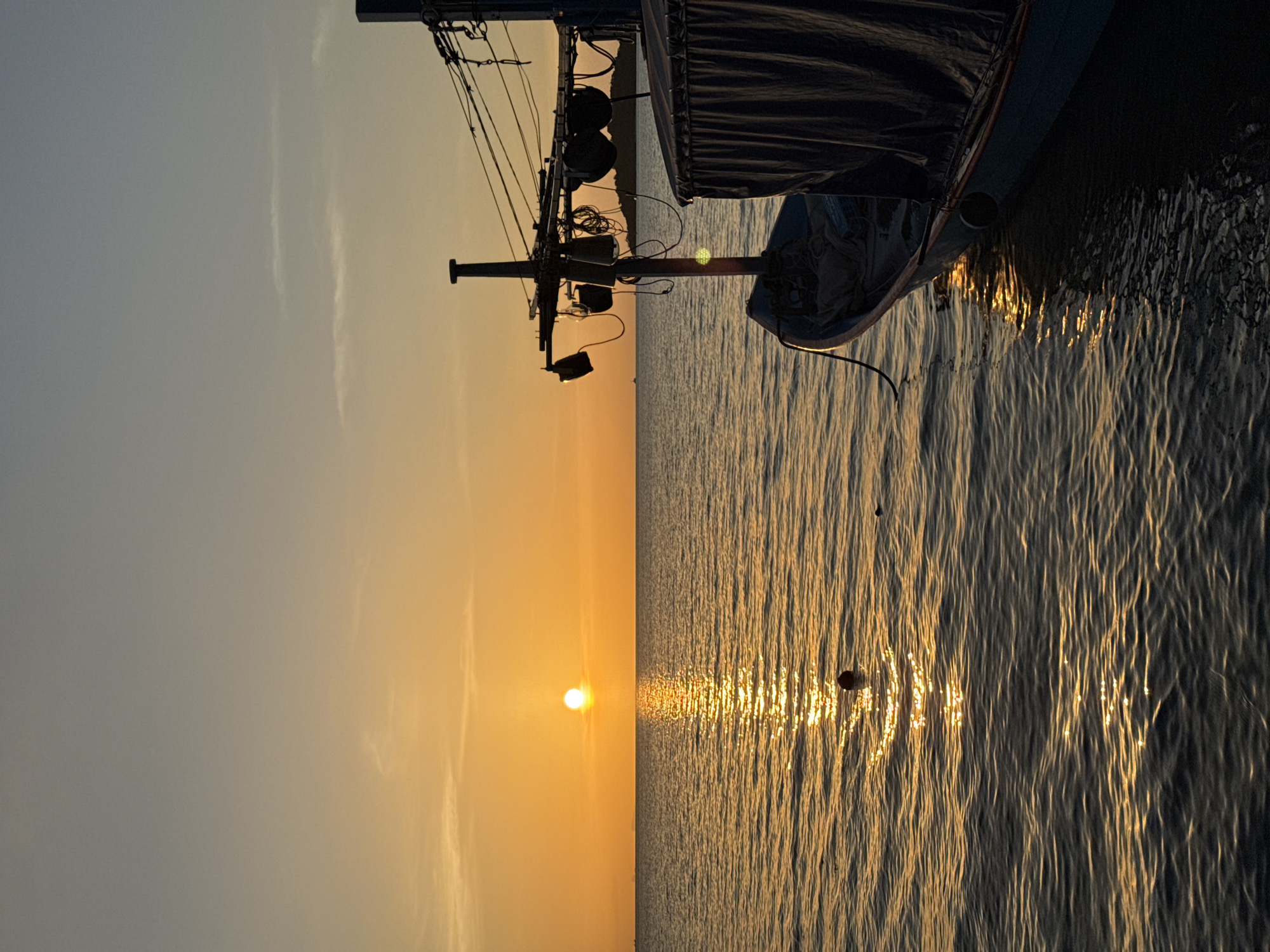 A fishing boat is moored in the calm ocean waters off the coast of Ko Samui District, Thailand, during a sunset. The sun, a bright orb, is positioned slightly left of center on the horizon, casting a golden path across the water. The sky transitions from a pale blue at the top to a warm orange glow near the horizon. The boat, silhouetted against the vibrant sky, has a blue hull and is partially covered with a dark tarp. A tall mast with rigging and various lights extends upwards from the boat. The water is gently rippled, reflecting the warm light of the setting sun. In the distance, a faint silhouette of land is visible on the horizon.