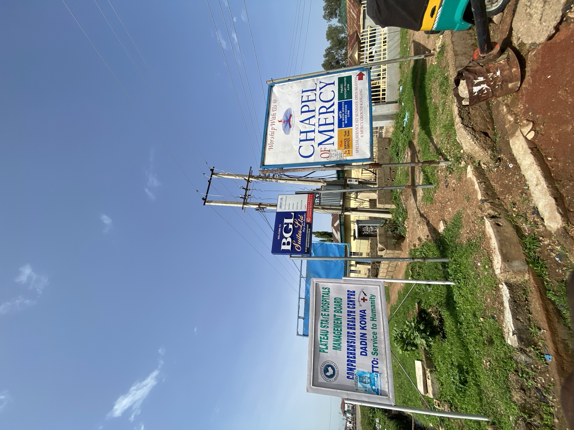 A street-level, eye-level, outdoor, daytime, long shot captures a bright, clear sky with sparse clouds above a street in Gura Top, Nigeria. Three signs stand prominently. The leftmost sign, a white rectangular board with a blue border, advertises the "PLATEAU STATE HOSPITALS MANAGEMENT BOARD COMPREHENSIVE HEALTH CENTRE DADIN KOWA" with the motto "Service to Humanity." A blue and white poster is affixed to the lower left corner of this sign. To the right of this, a smaller, dark blue rectangular sign with white lettering reads "BGL Suites Ltd" and lists various amenities such as "Executive Accommodation," "Restaurant," "Lounge Bar," "Gymnasium," and "Business Centre." Below this, contact numbers are listed. The largest sign, a white rectangular board with a blue border, is positioned furthest to the right. It displays "Worship With Us @" at the top, followed by the prominent text "CHAPEL OF MERCY." Below this, service times are listed for Sunday, Wednesday, and Thursday. At the bottom, it reads "SPECIAL SERVICE 1ST SUNDAYS: OPEN HEAVENS @ MERCY GROUND KANGANG."
The signs are supported by metal poles and are surrounded by overgrown grass and dirt. A portion of a yellow and green motorized tricycle is visible on the far right of the image. Electrical wires are strung across the top of the frame, supported by wooden poles and transformers. In the background, houses with corrugated iron roofs and a fence can be seen. The overall impression is a functional street scene with commercial and religious establishments.